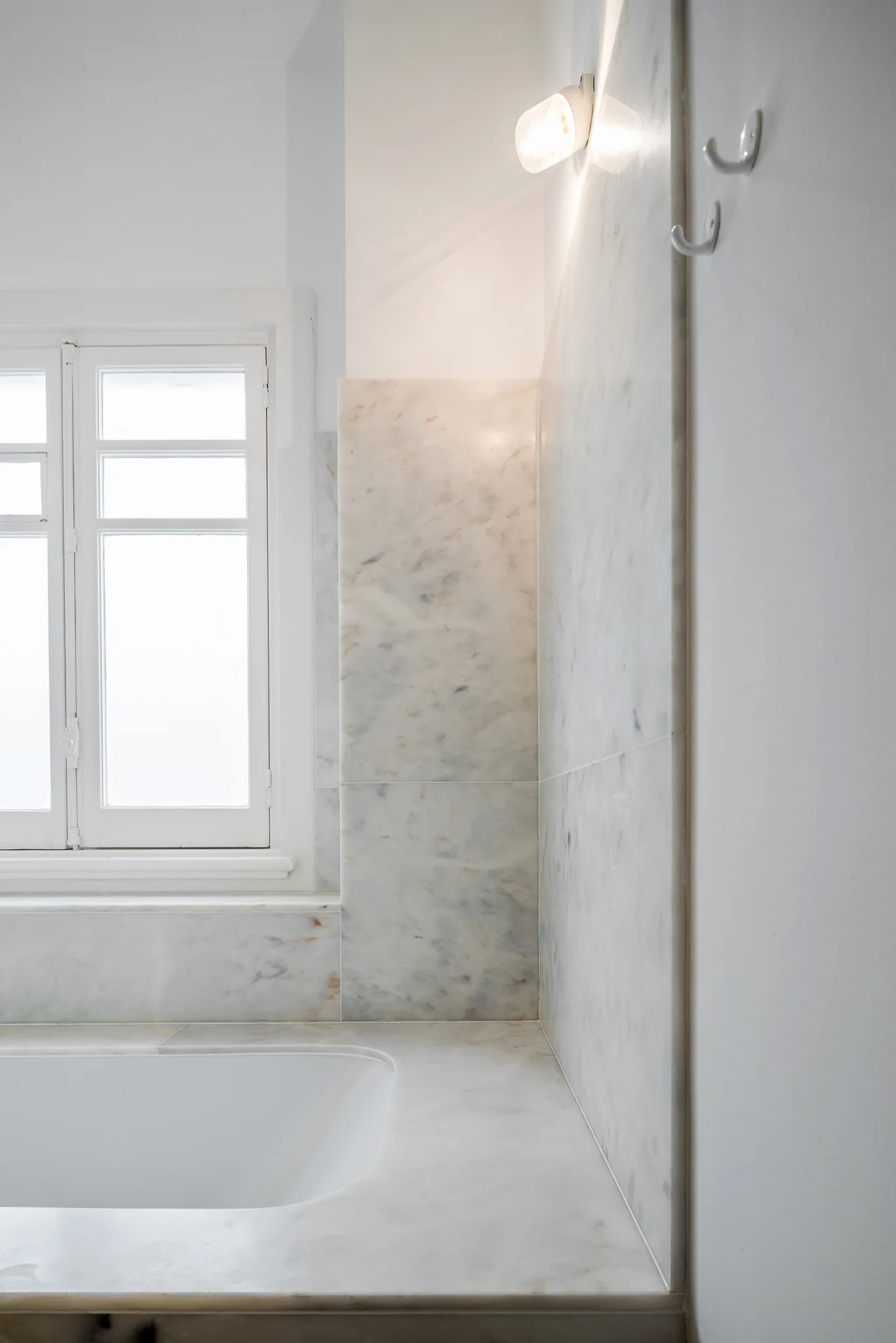 A polished white marble bathtub deck and wall surround next to a classic white framed window with a small wall light.