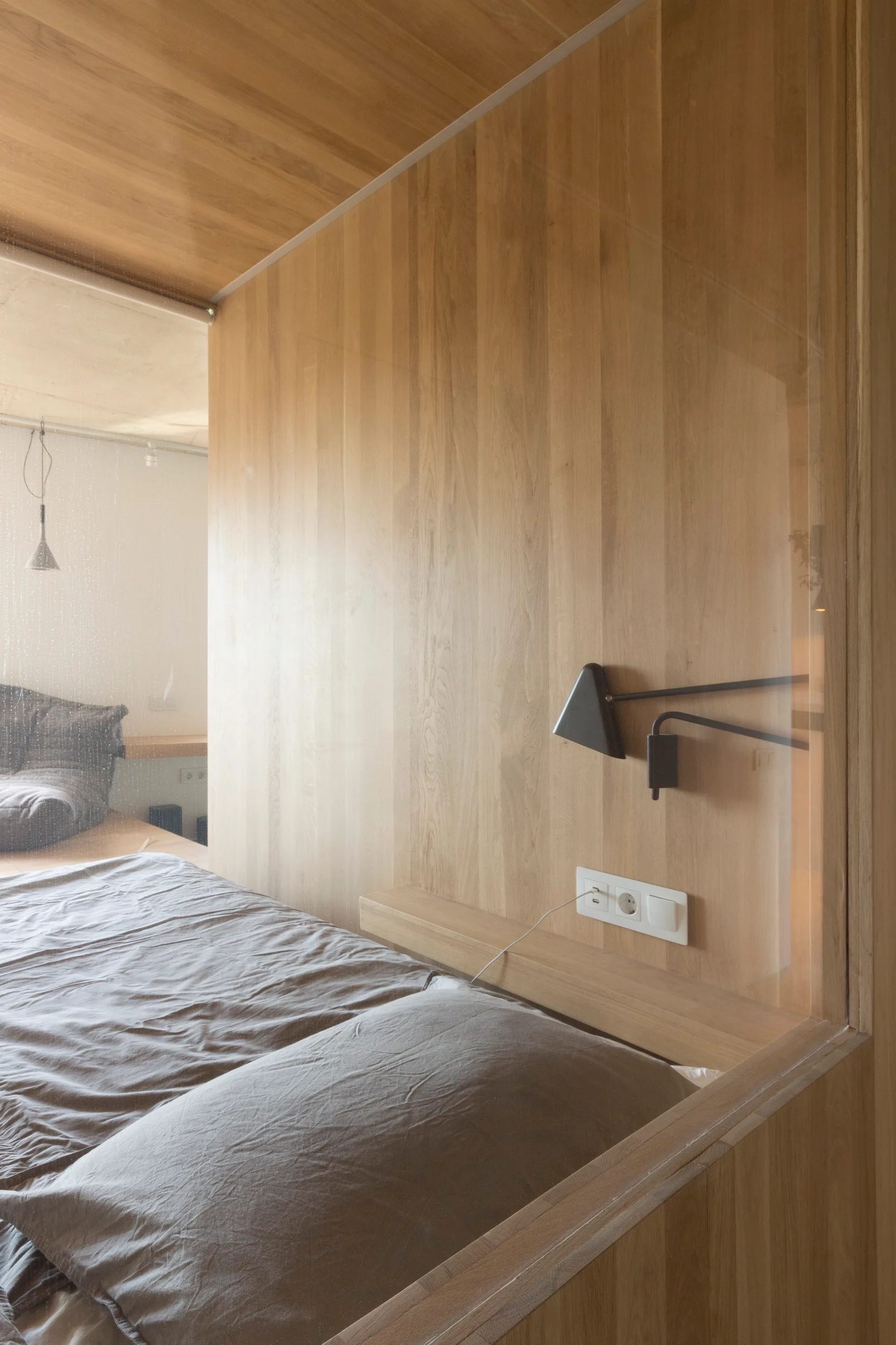 A minimalist bedroom features a bed with grey sheets against a vertical wood-paneled wall next to a clear glass partition and a black sconce.
