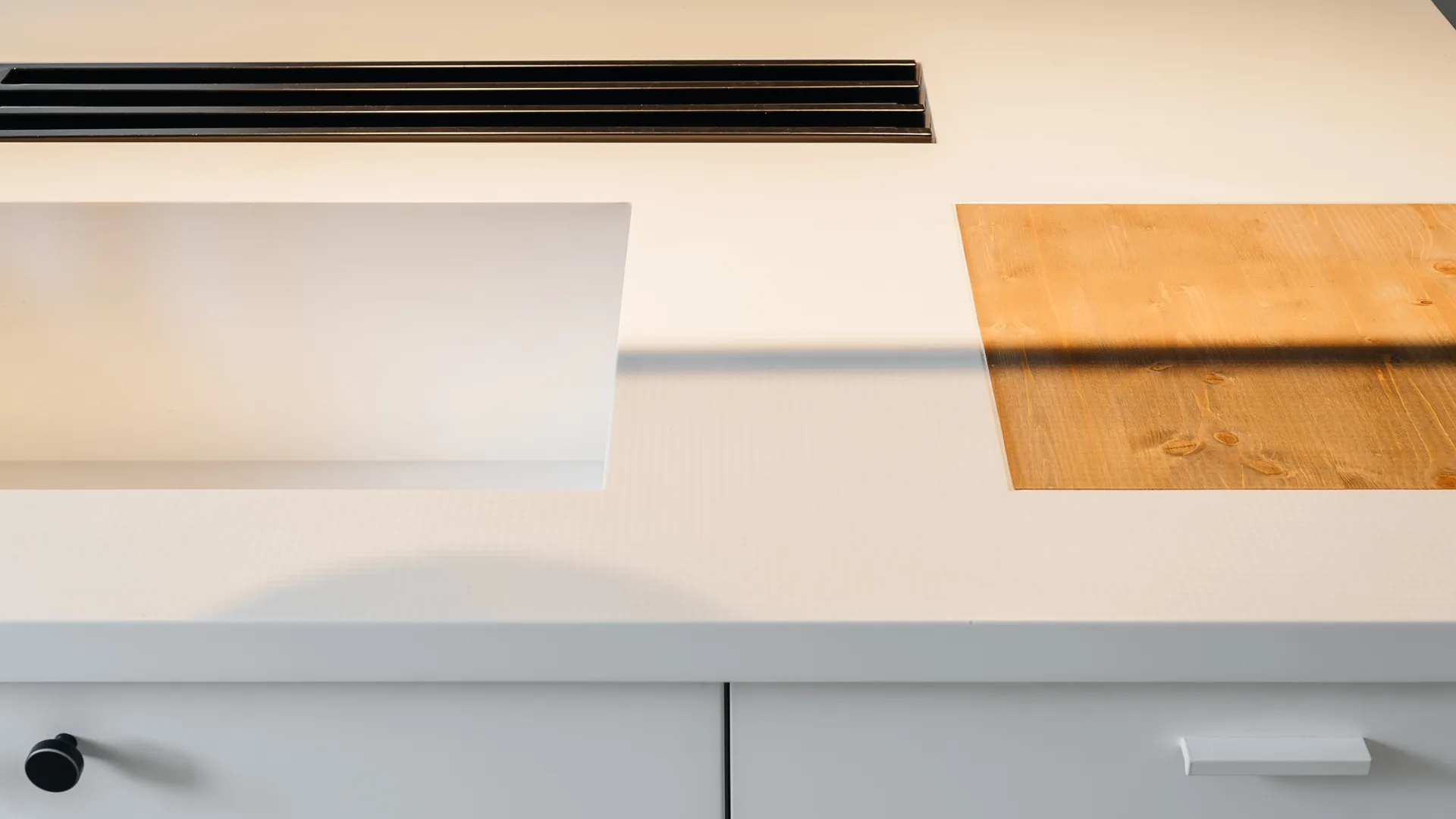 A white kitchen countertop features an integrated sink basin next to an inset wooden cutting board and a black stove vent.