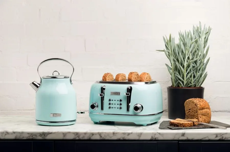 A light blue kettle and matching toaster sit on a veined marble countertop next to a potted plant and sliced bread against a white brick wall.