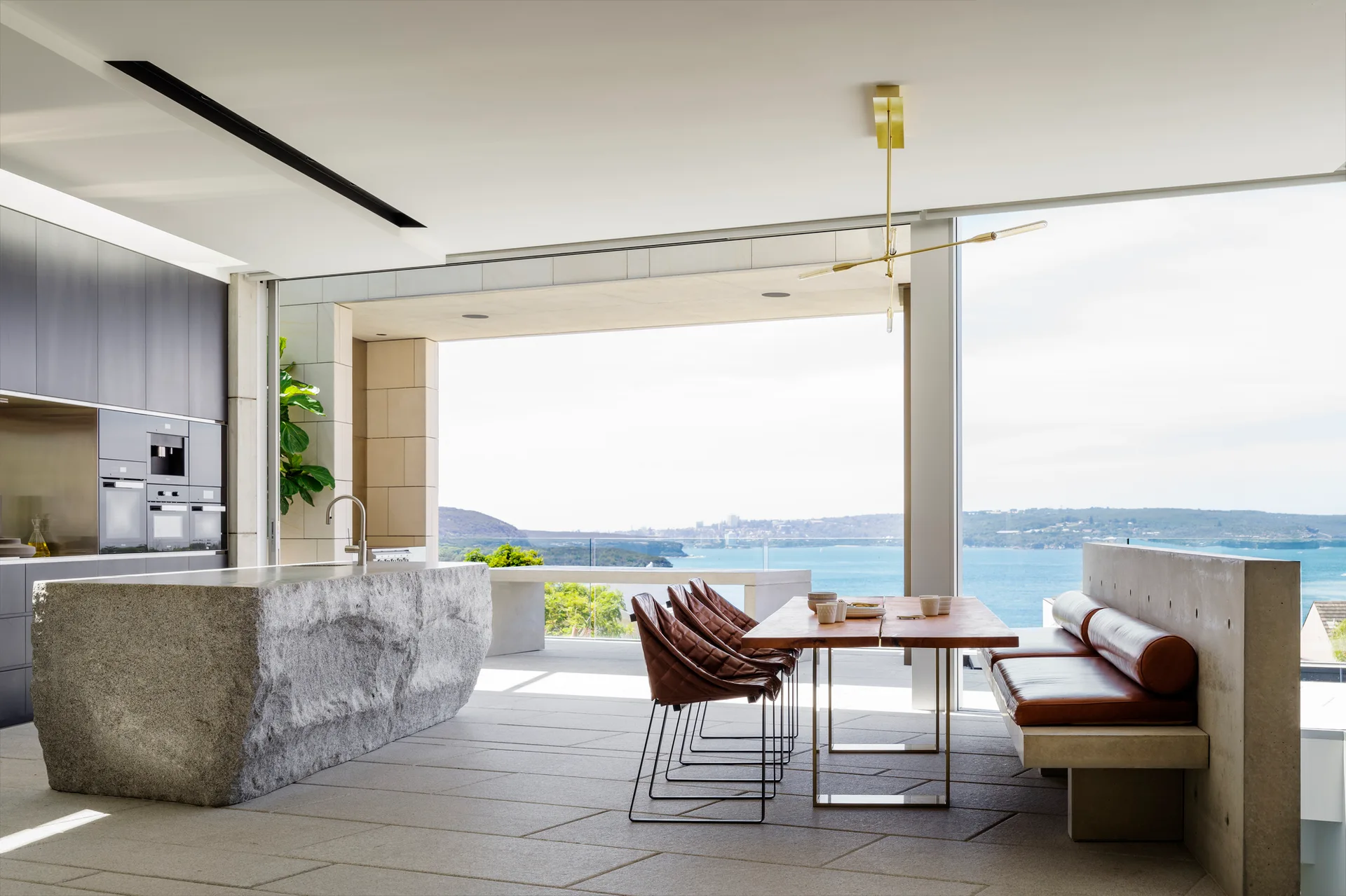 A modern open-plan kitchen with a large rough-hewn stone island, wooden dining table, and leather-cushioned concrete bench overlooking the ocean.