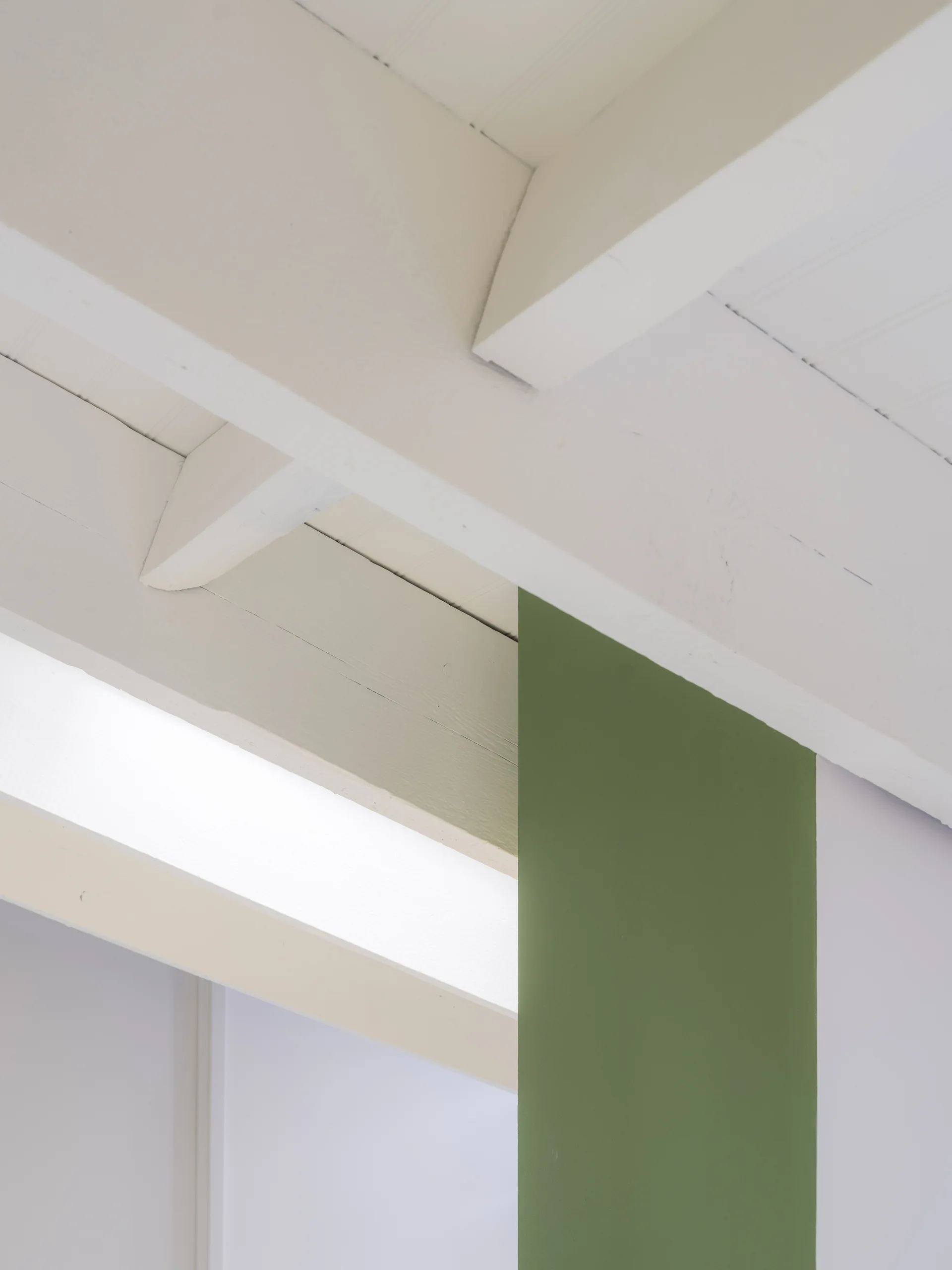 A close-up view of exposed wooden ceiling beams painted white next to a single vertical structural column painted matte olive green.