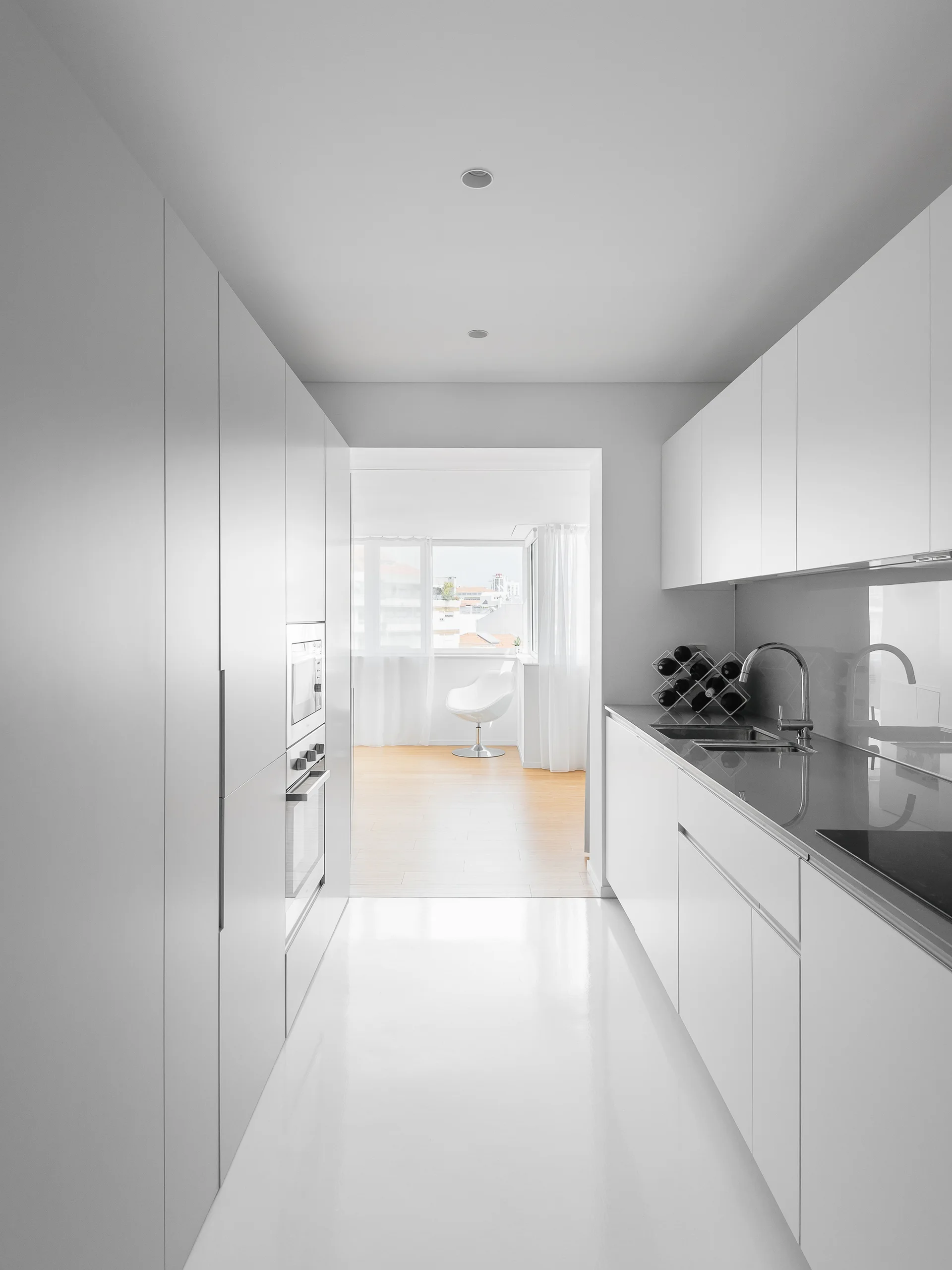 A minimalist white galley kitchen features handleless cabinetry, a gray countertop with a steel faucet, and a highly reflective white glossy floor.