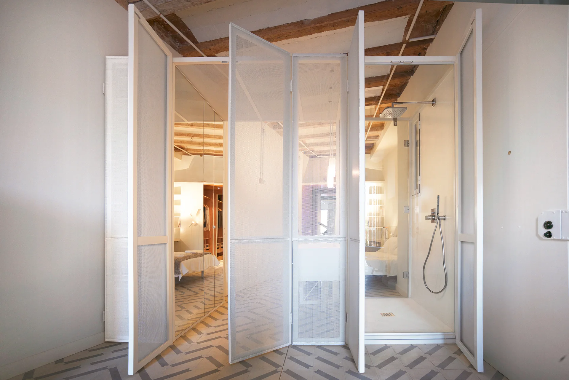 A modern bedroom and bathroom space divided by a white perforated metal mesh partition, featuring chevron tiled floors and exposed ceiling beams.