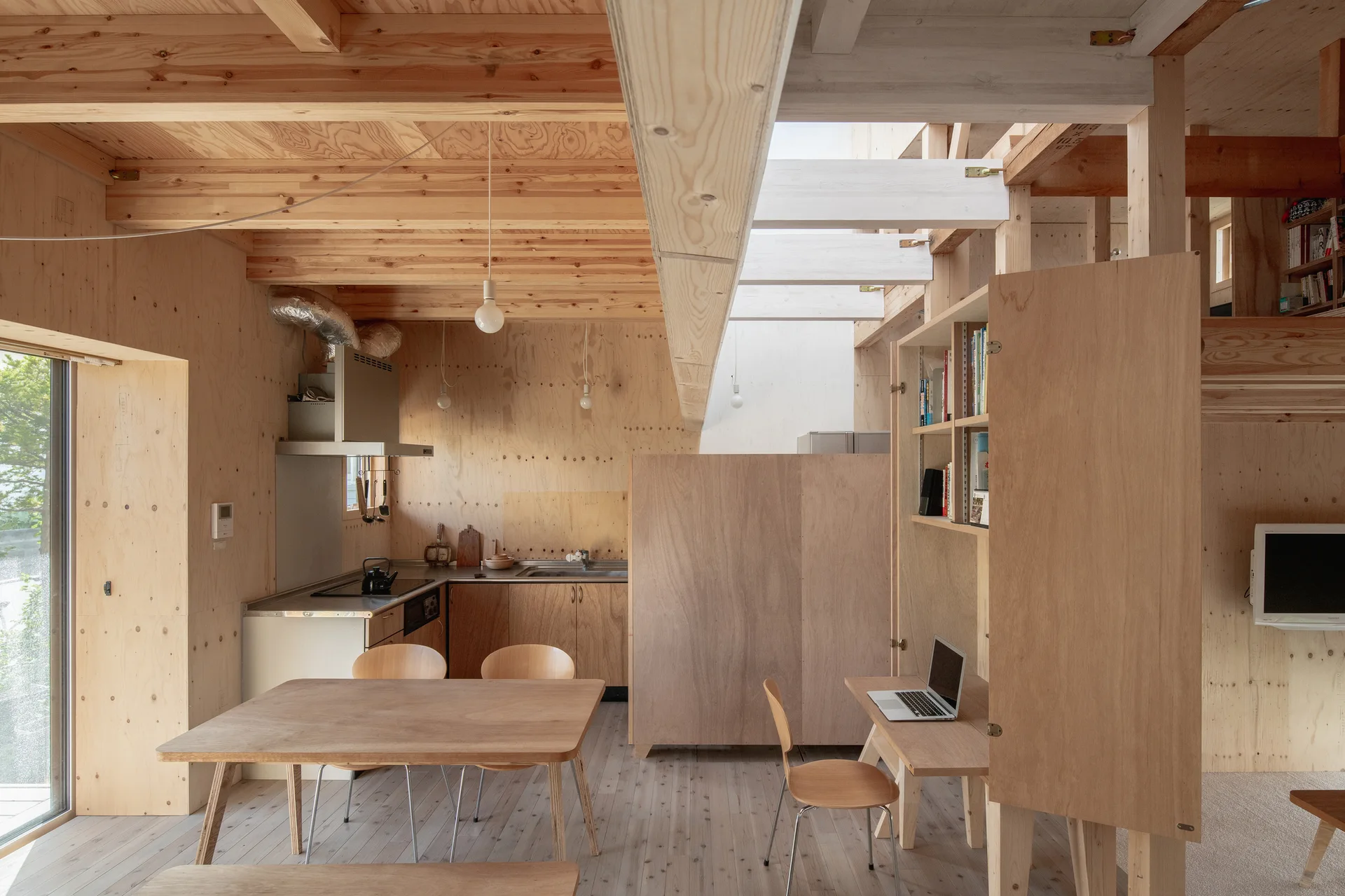 An open-concept room features raw plywood walls, a small wooden dining set, an integrated study desk, and exposed ceiling joists.