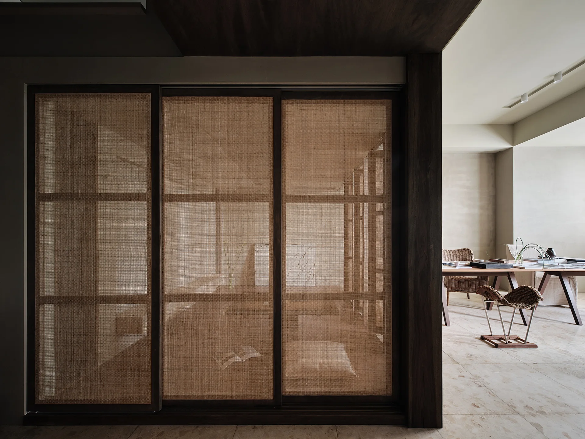 A dark wood sliding screen with woven cane panels partitions a minimalist interior featuring large stone floor tiles and a workspace in the background.
