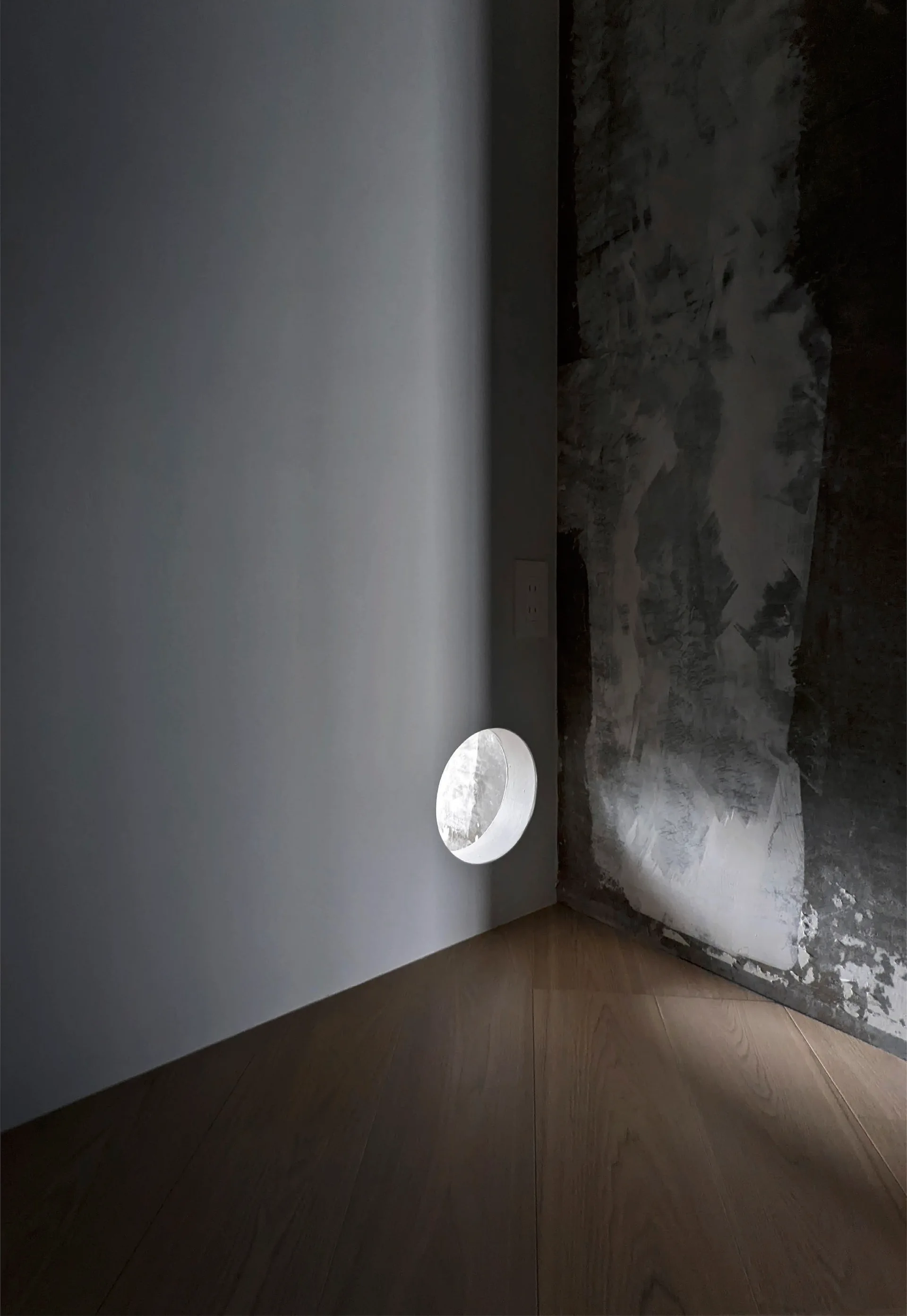 A minimalist interior corner features a smooth white wall with a circular cutout meeting a textured grey plaster wall over wood flooring.