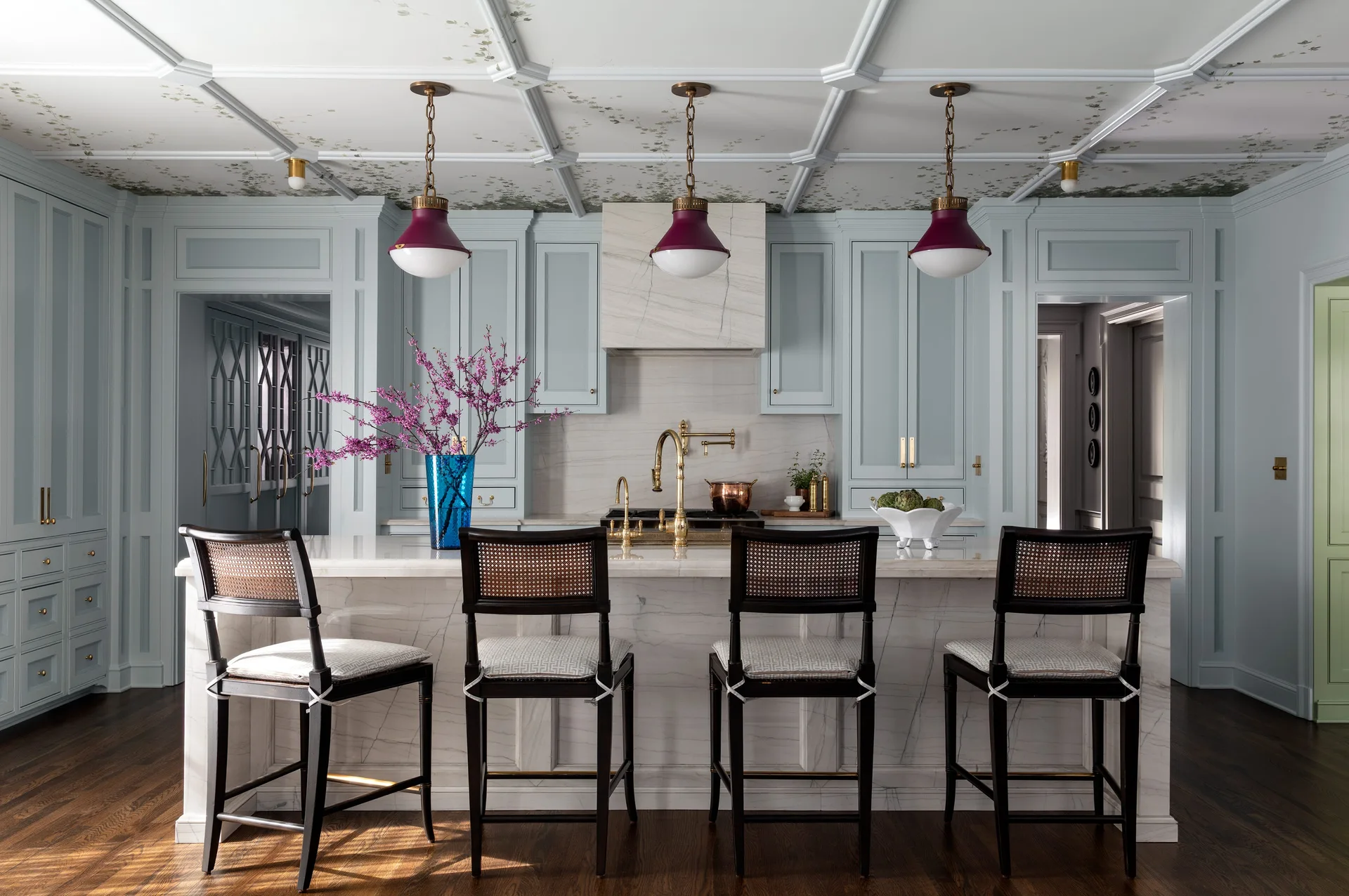 A light blue kitchen features a marble-topped island with four dark wood stools beneath a white coffered ceiling decorated with botanical wallpaper.