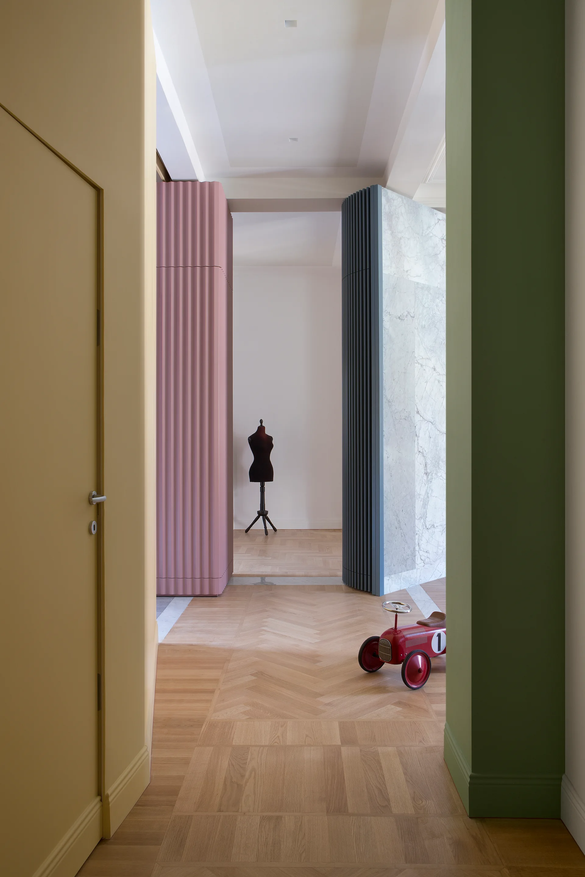 A colorful hallway featuring blue, pink, green, and yellow architectural pillars, herringbone hardwood floors, and a red toy car.