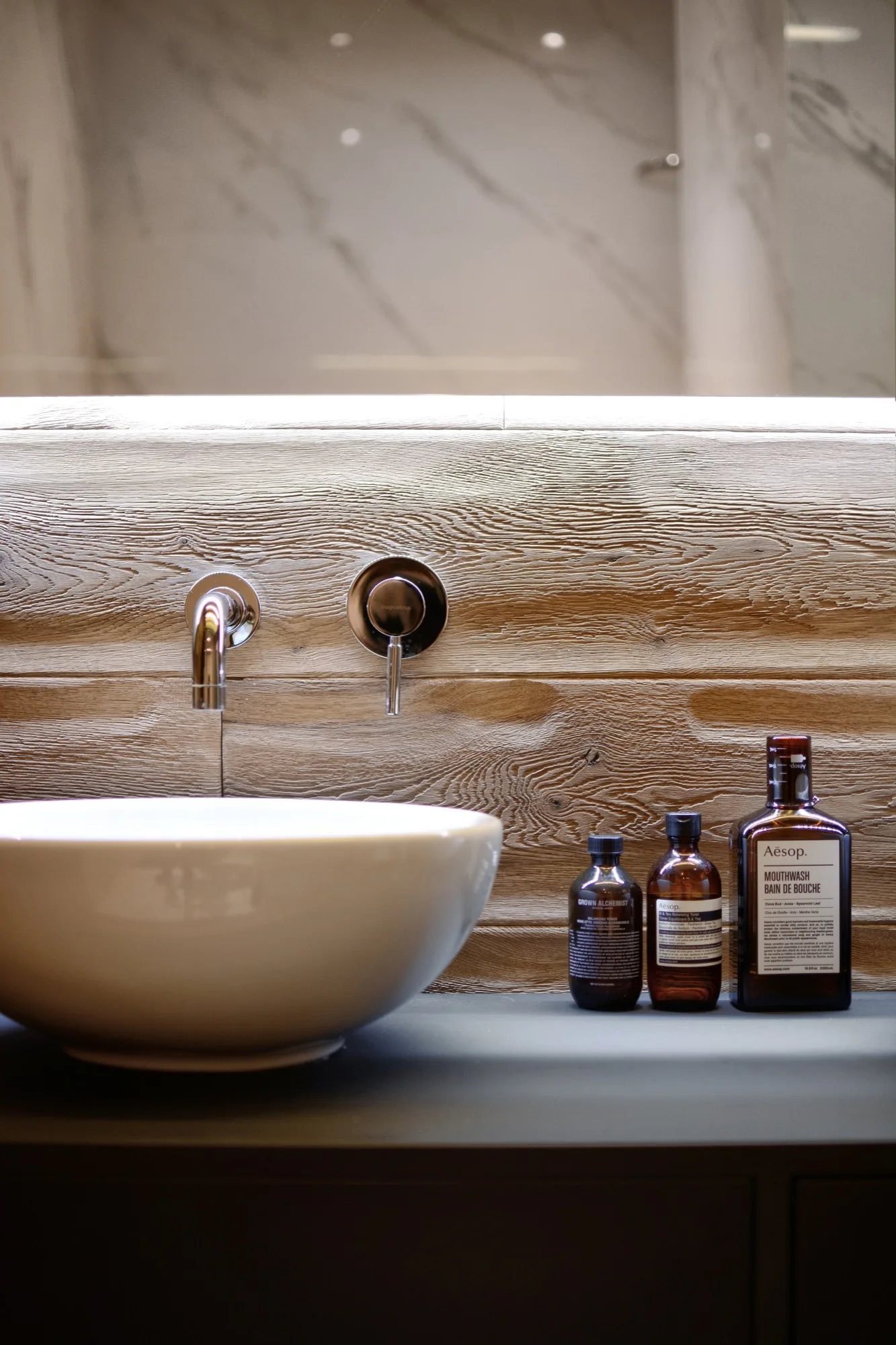 A white ceramic vessel sink sits on a dark vanity counter against a textured wood-grain wall with a wall-mounted chrome faucet and amber glass bottles.