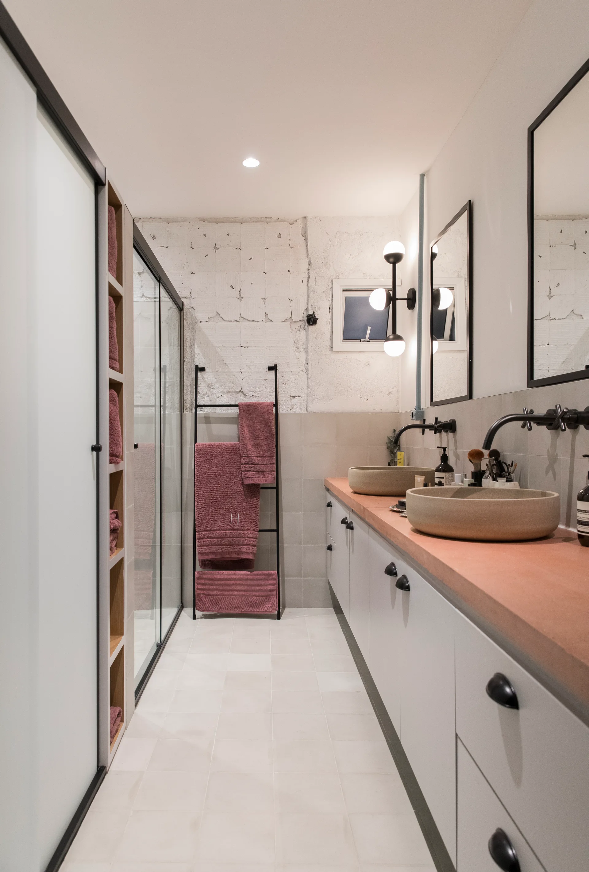 A modern narrow bathroom with a peach-colored double vanity, round stone vessel sinks, black fixtures, and a glass shower enclosure next to a towel ladder.
