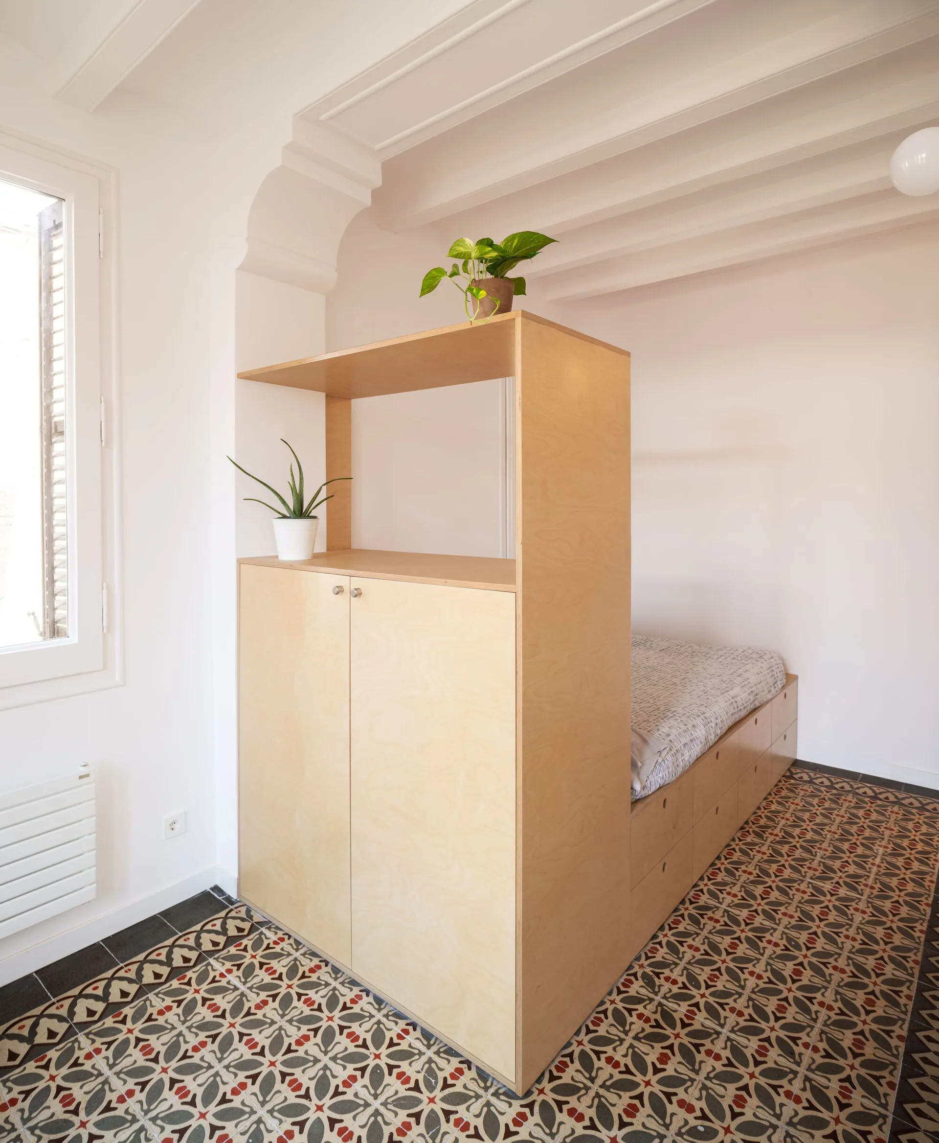 A minimalist bedroom featuring a custom light wood plywood platform bed with integrated cabinet storage on a patterned tile floor.