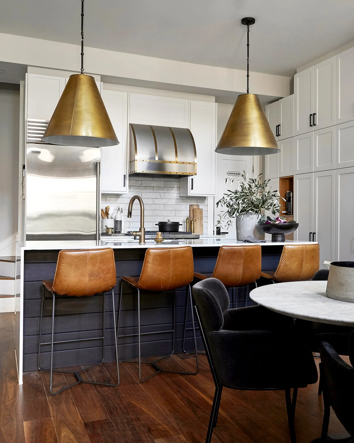 A modern kitchen features a navy blue island with leather bar stools, white shaker cabinetry, and two large brass pendant lights.