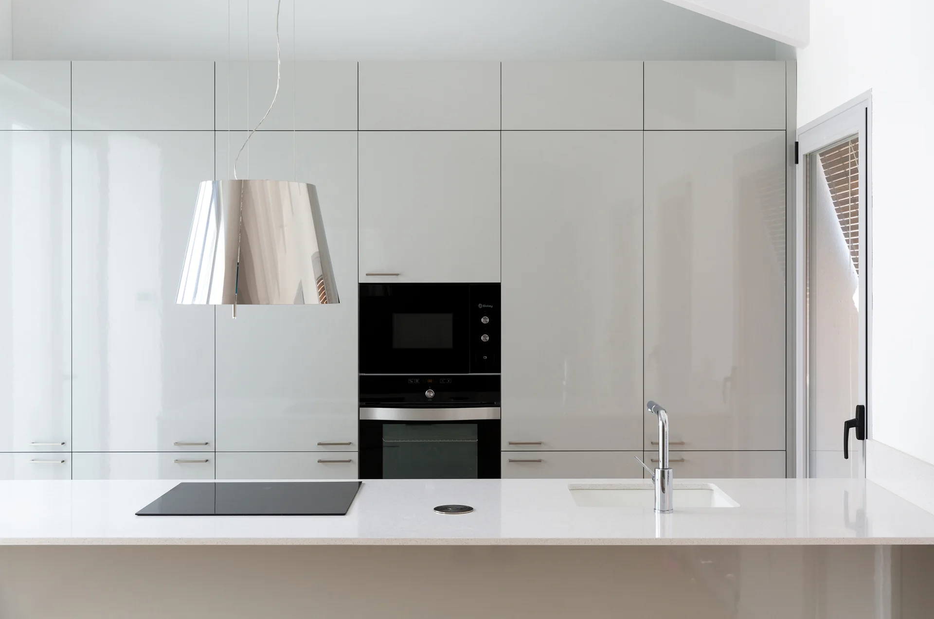 A minimalist kitchen features high-gloss light grey cabinets, a white island with a cooktop and sink, and a metallic conical pendant light.