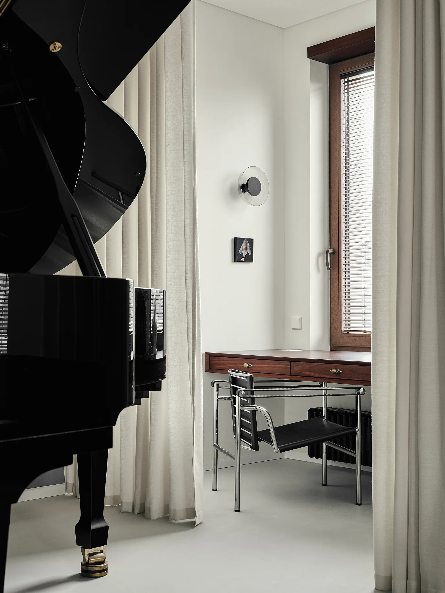 A black grand piano stands near a wooden writing desk with a chrome and leather chair by a window with white curtains.