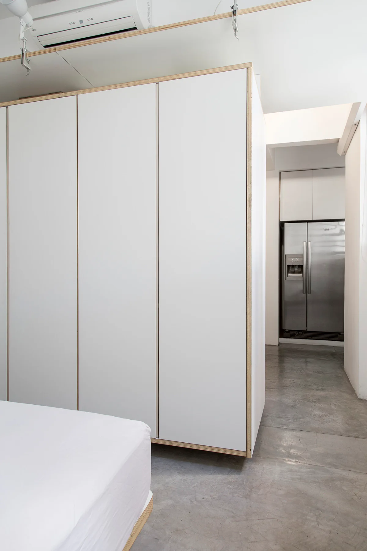 A minimalist bedroom featuring a large white wardrobe with exposed plywood edges, concrete flooring, and a view into a kitchen.