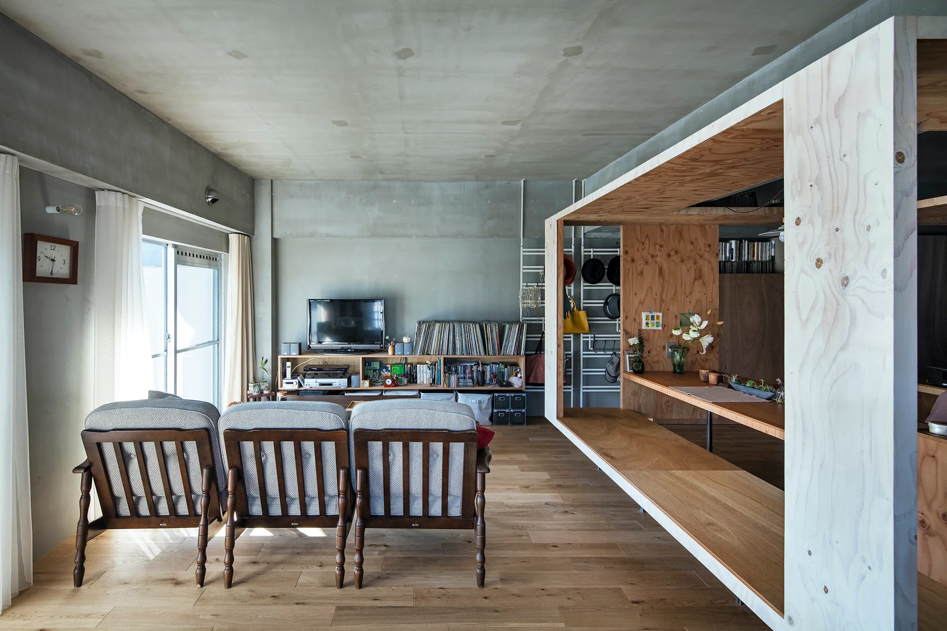 A minimalist living room features three grey armchairs, an open plywood partition structure, and exposed concrete walls and ceiling.