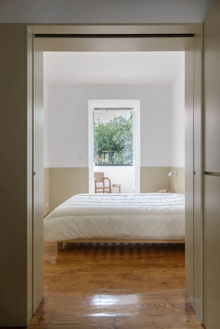 A view through a beige door frame into a minimalist bedroom featuring white and beige walls, a platform bed, glossy wood floors, and a window.