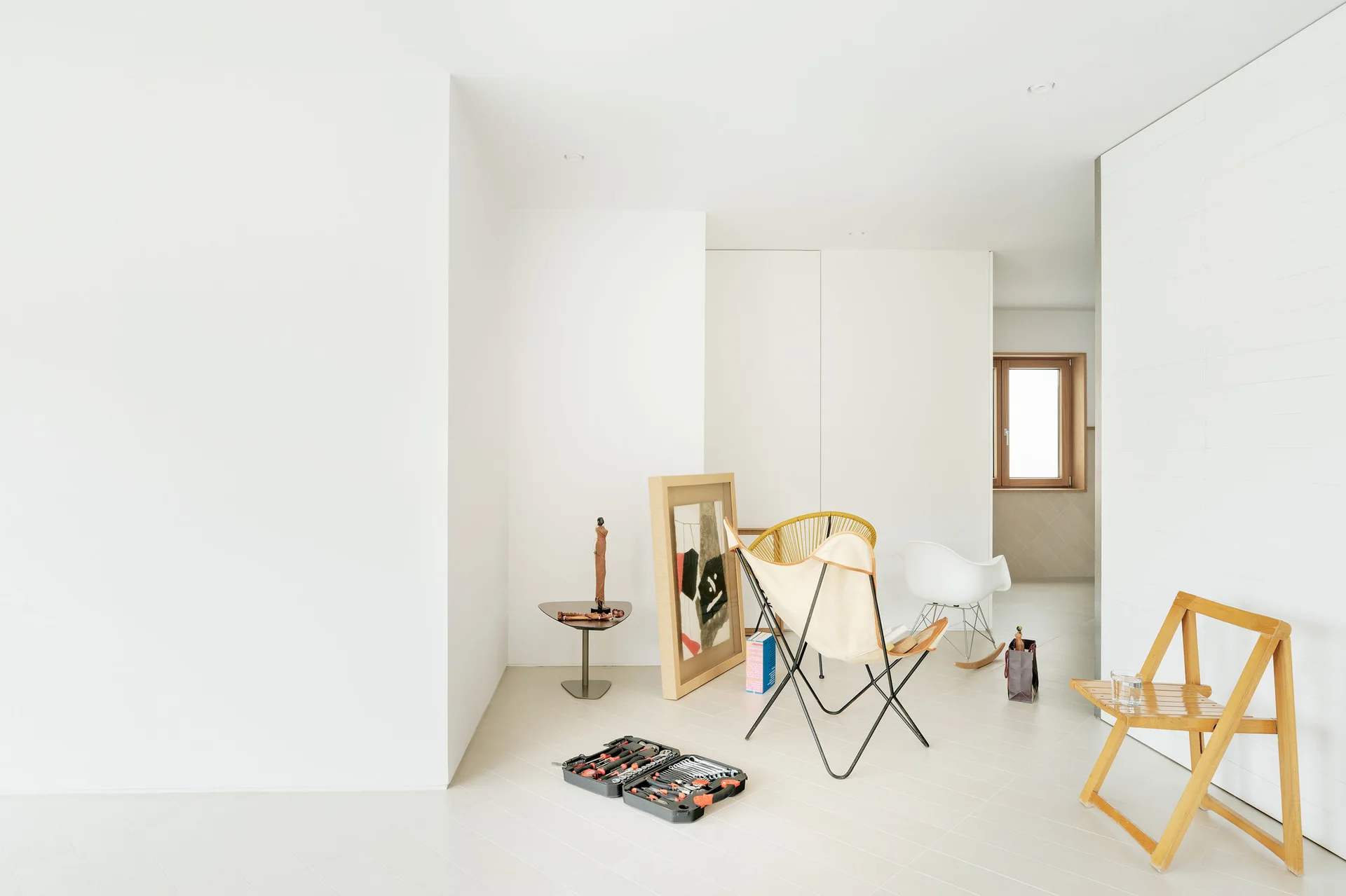 A minimalist white room features scattered mid-century chairs, leaning framed artwork, and light-toned floor tiles.