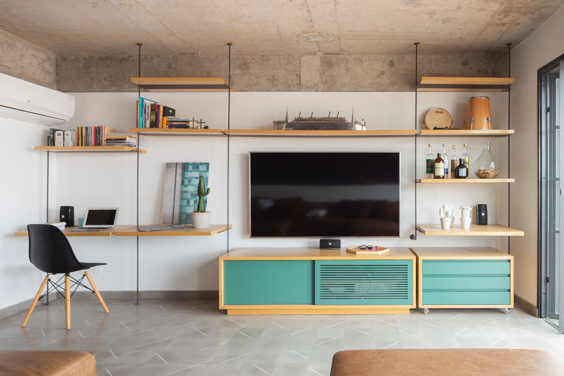 A wall-mounted wooden shelving system includes an integrated desk and television above low teal cabinets set against an exposed concrete ceiling.
