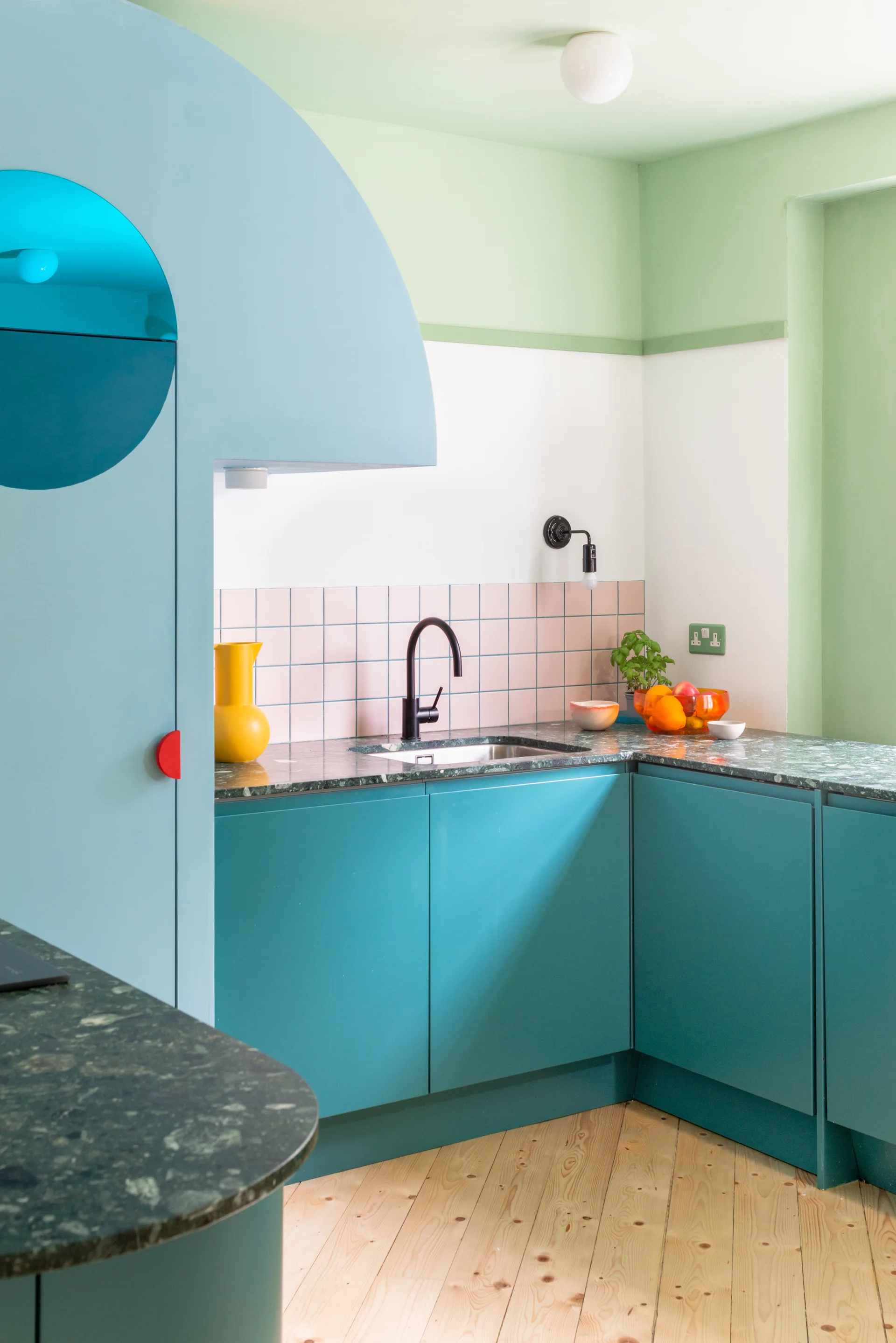 Teal kitchen cabinets with a green stone countertop, a pink square tile backsplash, and a black gooseneck faucet sit on light wood floors.