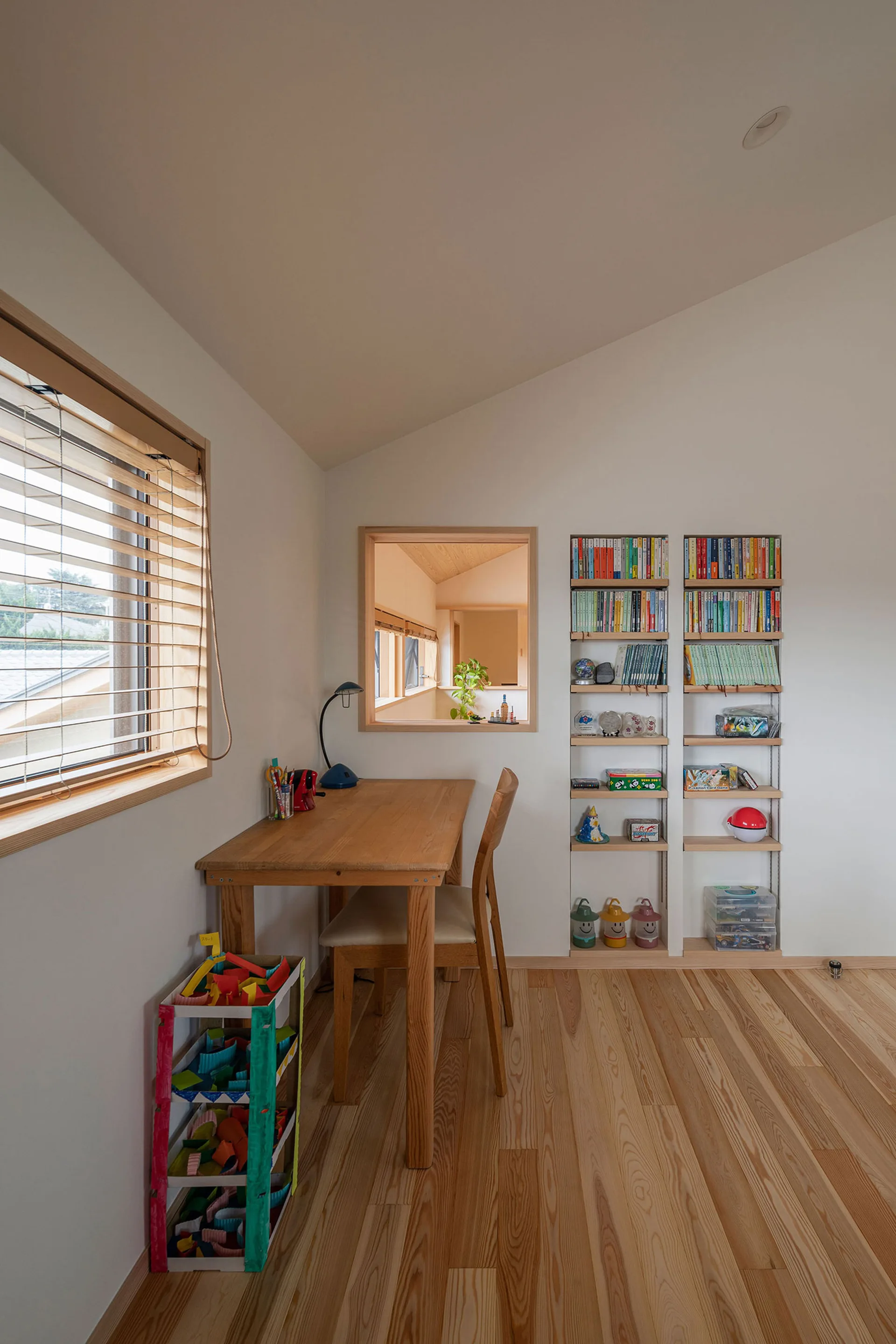 A minimalist children's study desk and chair sit next to a wall featuring a small interior cutout window and built-in recessed bookshelves.