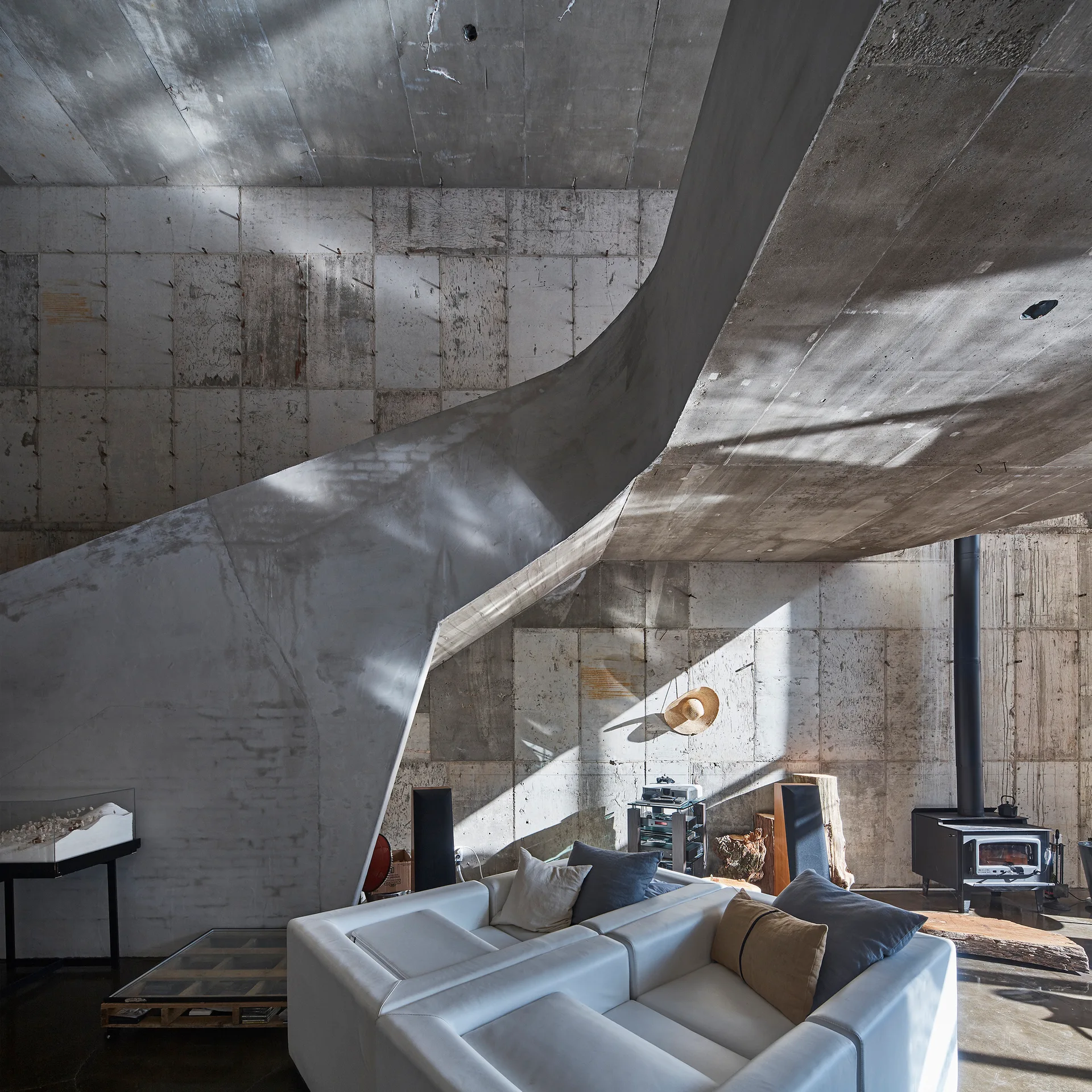 A low-profile white sofa sits below a massive curved concrete staircase in a brutalist living room with board-formed concrete walls.