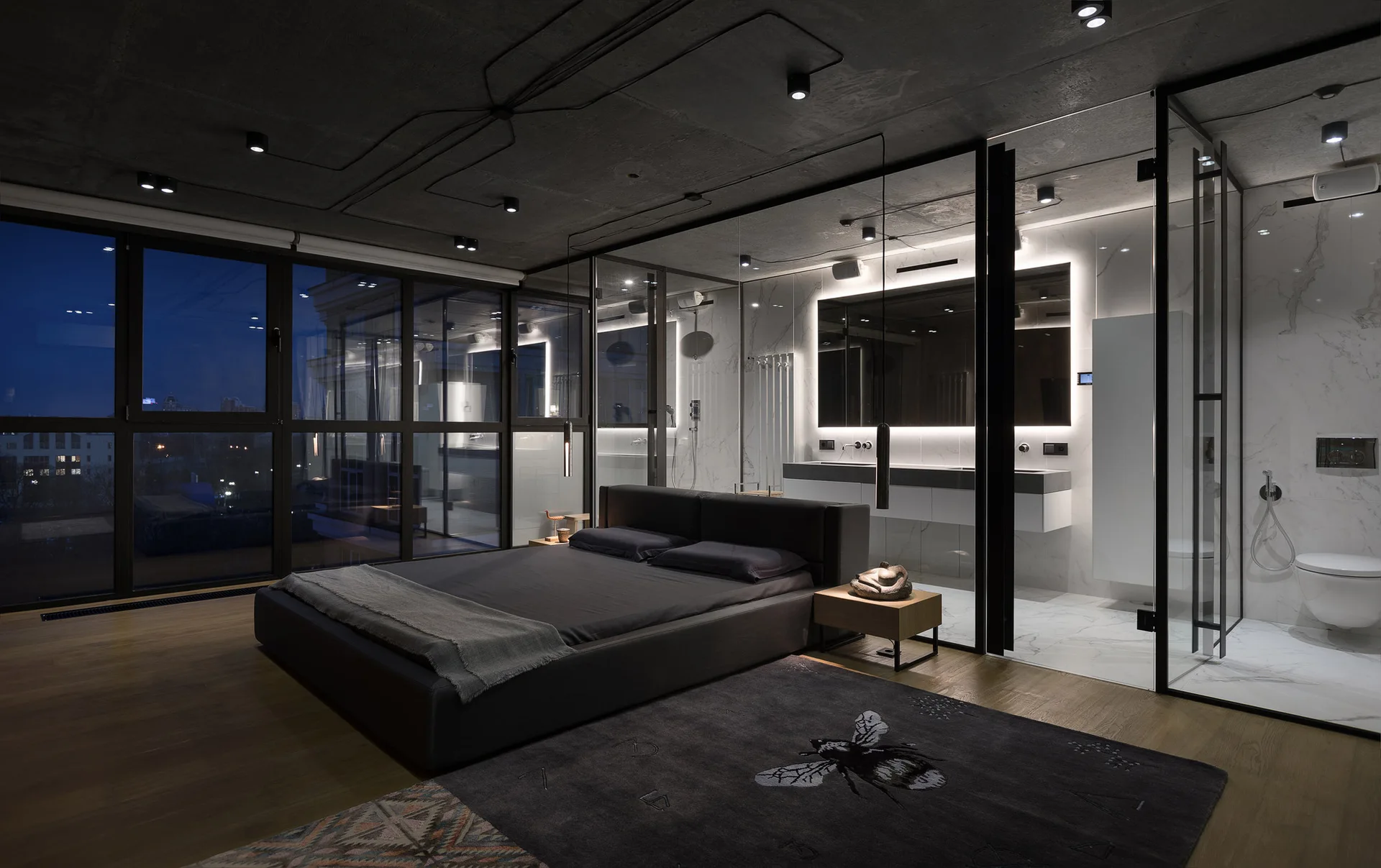 A modern bedroom features a dark upholstered bed next to a glass-walled bathroom with white marble tiling and an exposed concrete ceiling.