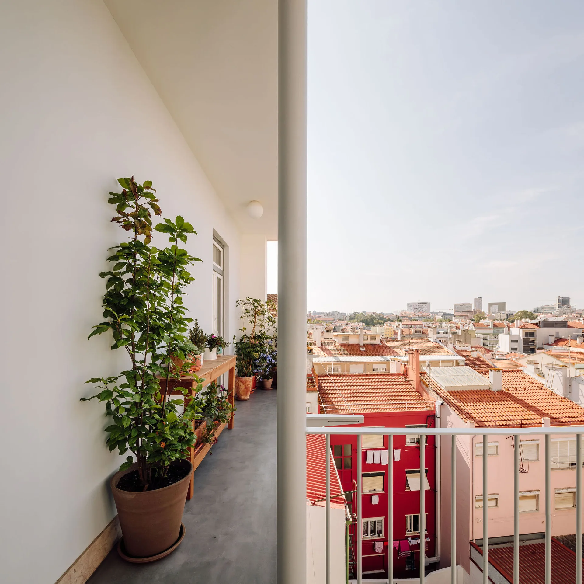 A narrow concrete balcony with a white railing features potted plants, a wooden console table, and a wide view of orange-tiled city rooftops.