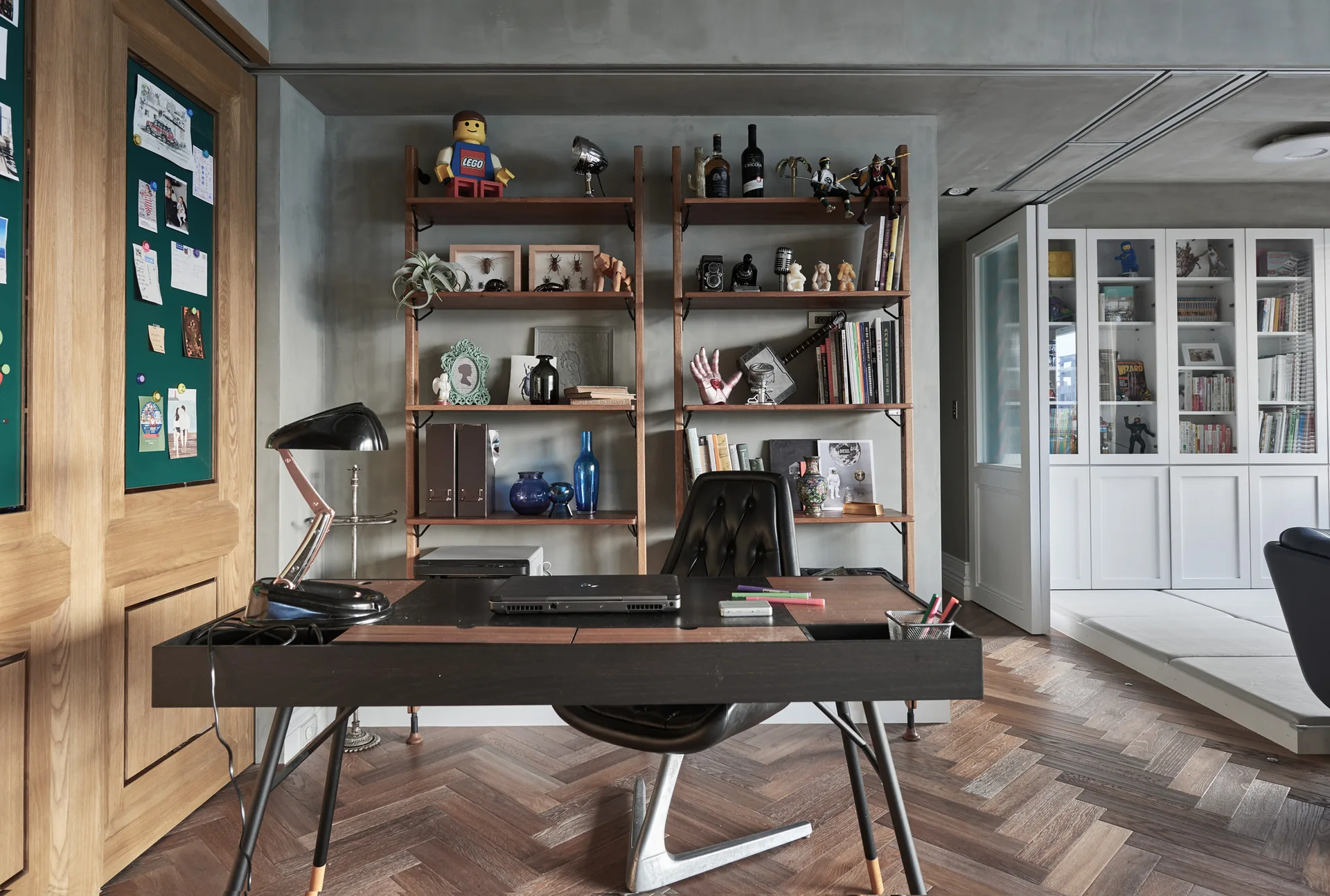 A home office featuring a dark wood desk, a tufted leather chair, and tall open wooden shelving against a textured grey concrete wall.
