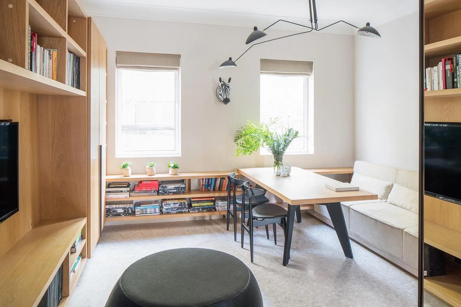 A bright dining room features a light wood table, a low bench sofa, matching built-in shelving, and a black branching pendant light.