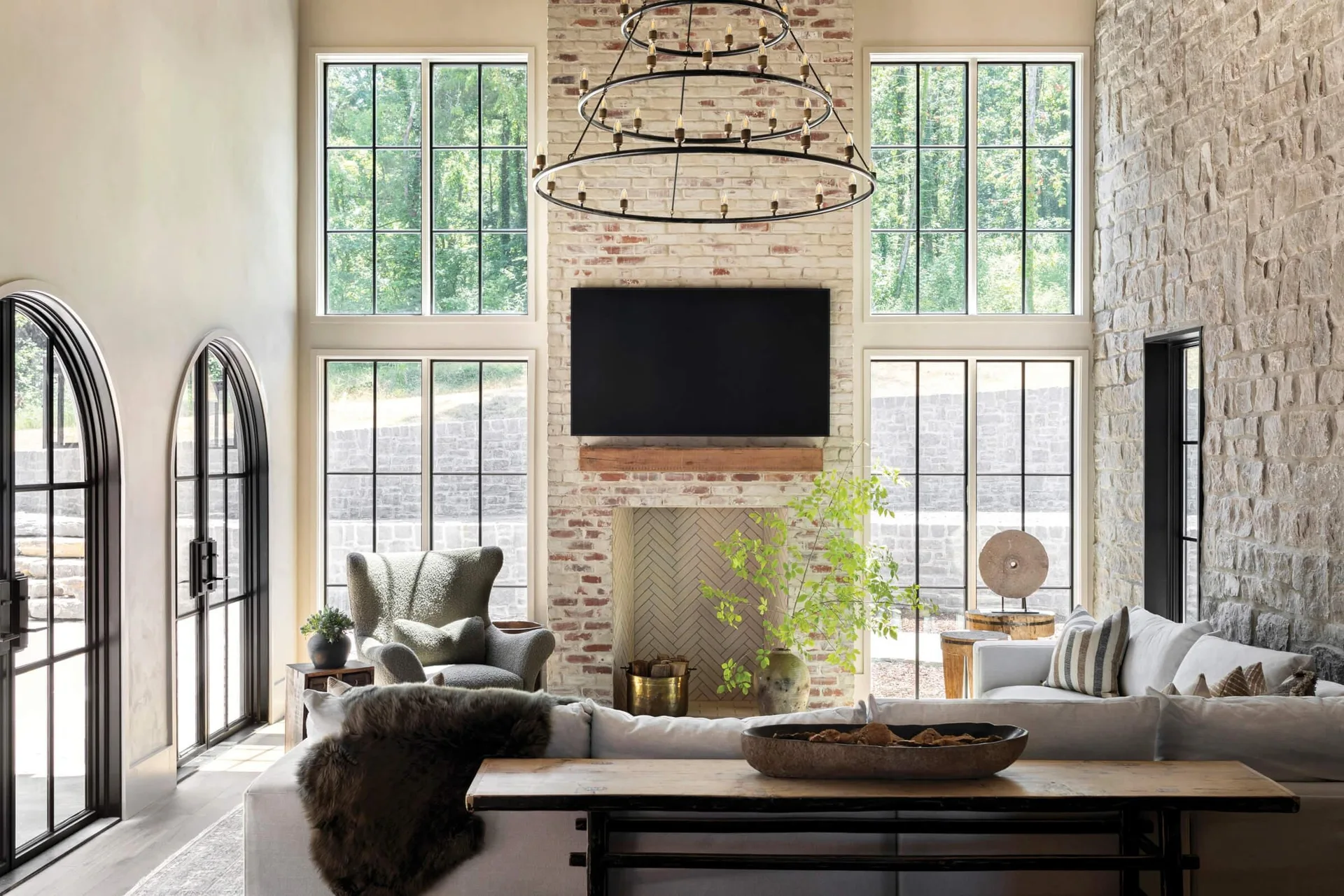 A high-ceilinged living room featuring a tall brick fireplace, a tiered metal chandelier, and a large sectional sofa flanked by gridded windows.