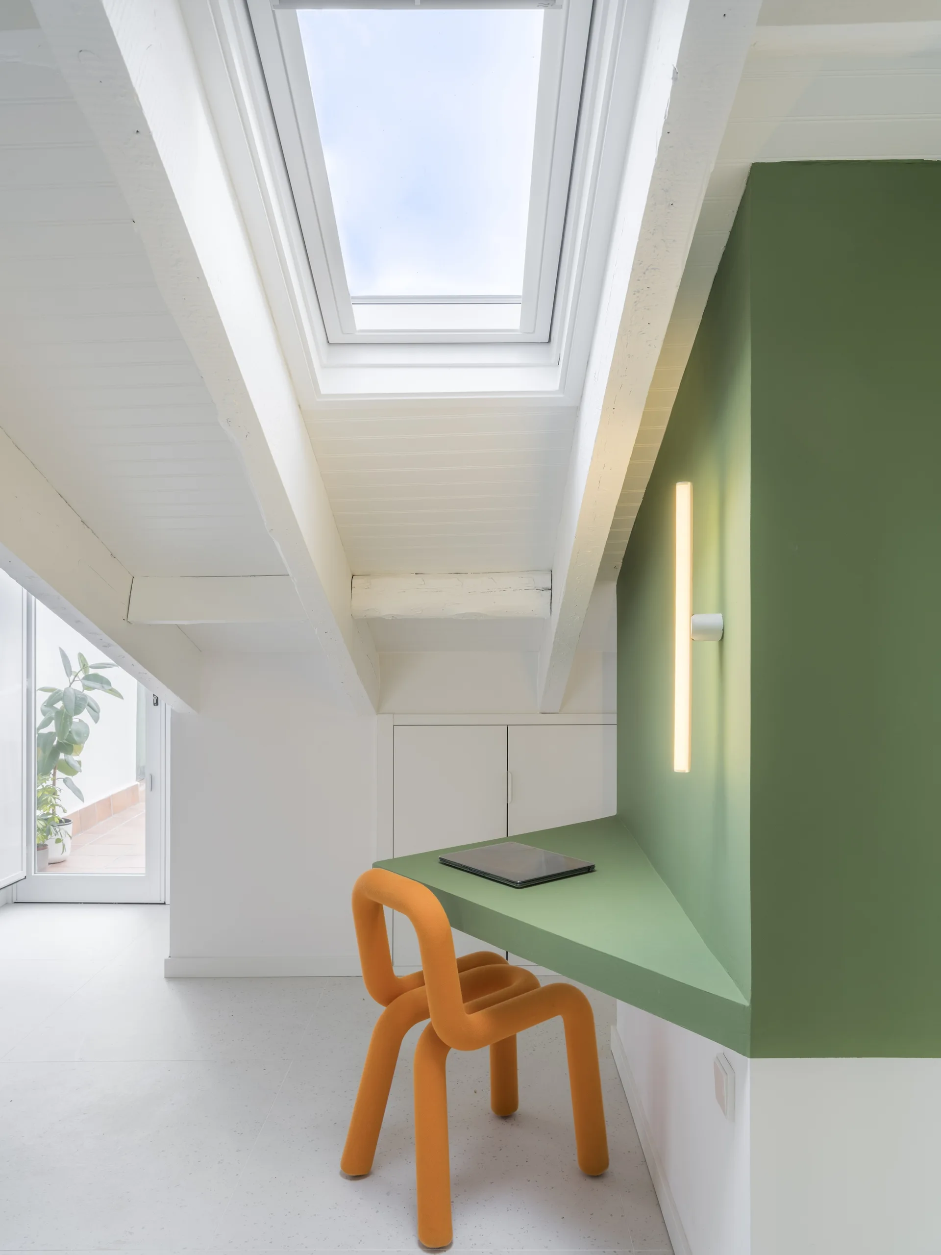 A modern attic home office features a built-in green corner desk, an orange tubular chair, and a skylight in the sloped white ceiling.