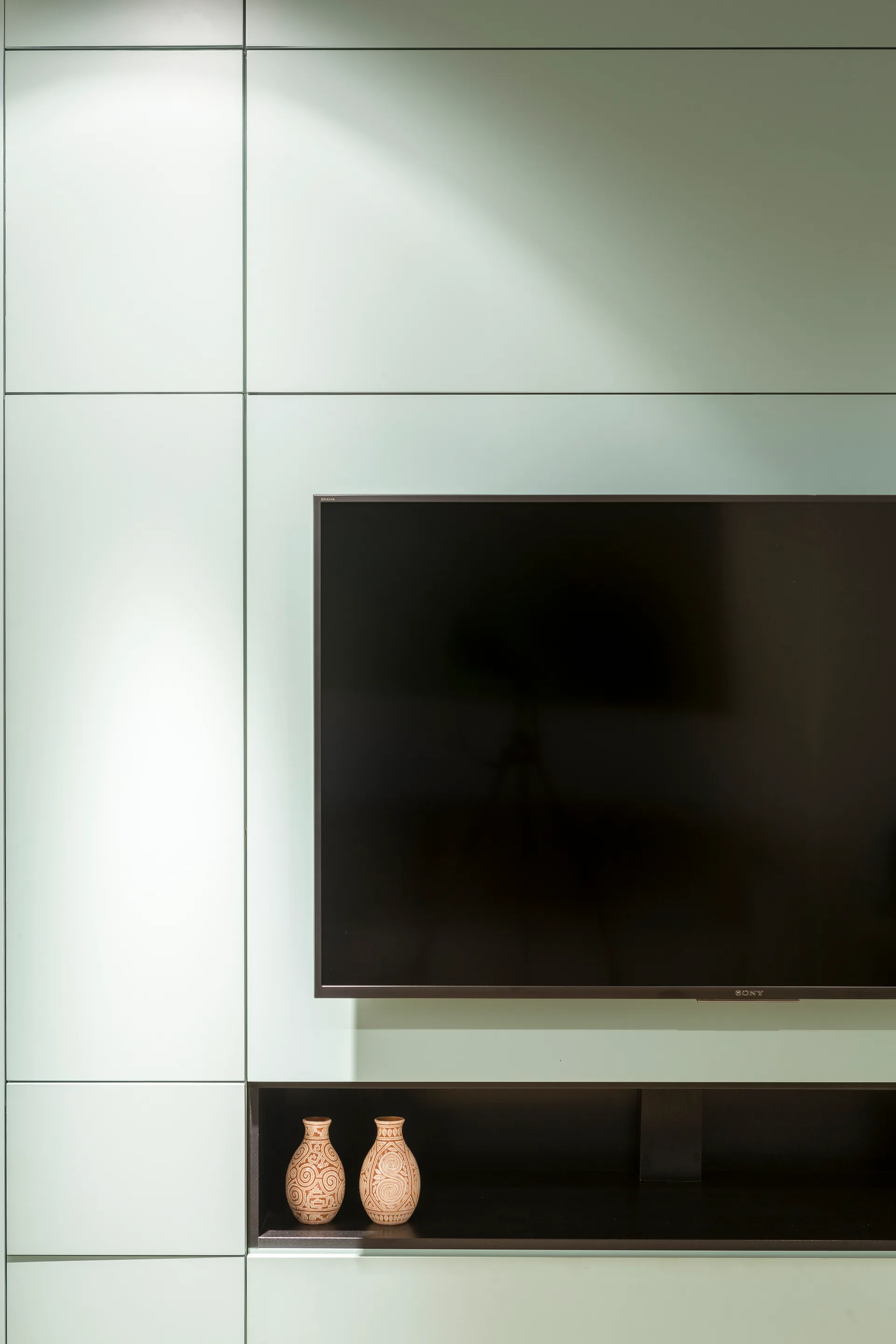 A flat-screen television is mounted on a mint green paneled wall above a dark recessed shelf containing two patterned terracotta vases.