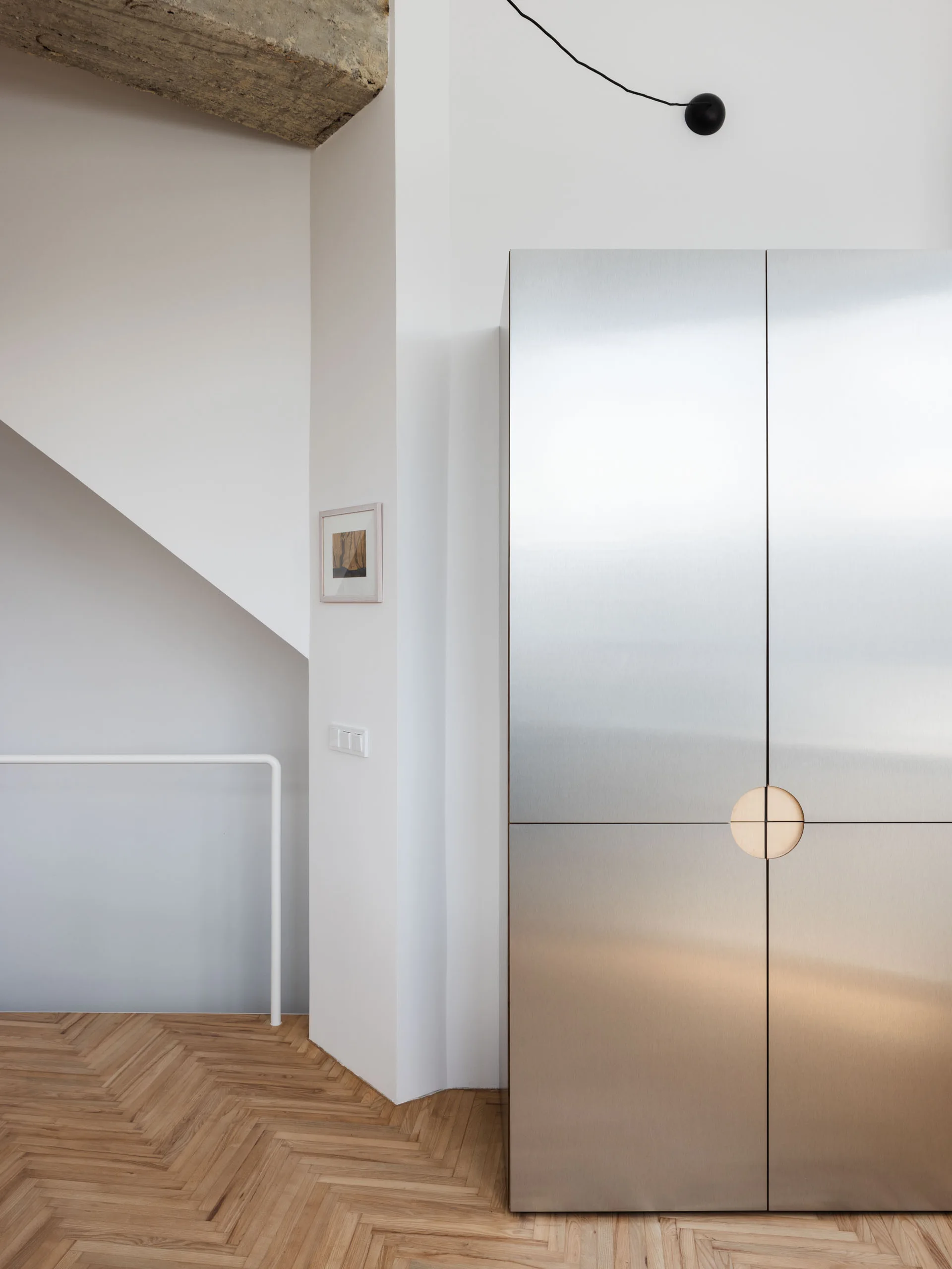A tall brushed metal cabinet with a circular cutout handle stands on herringbone wood floors next to a white hallway wall and staircase.