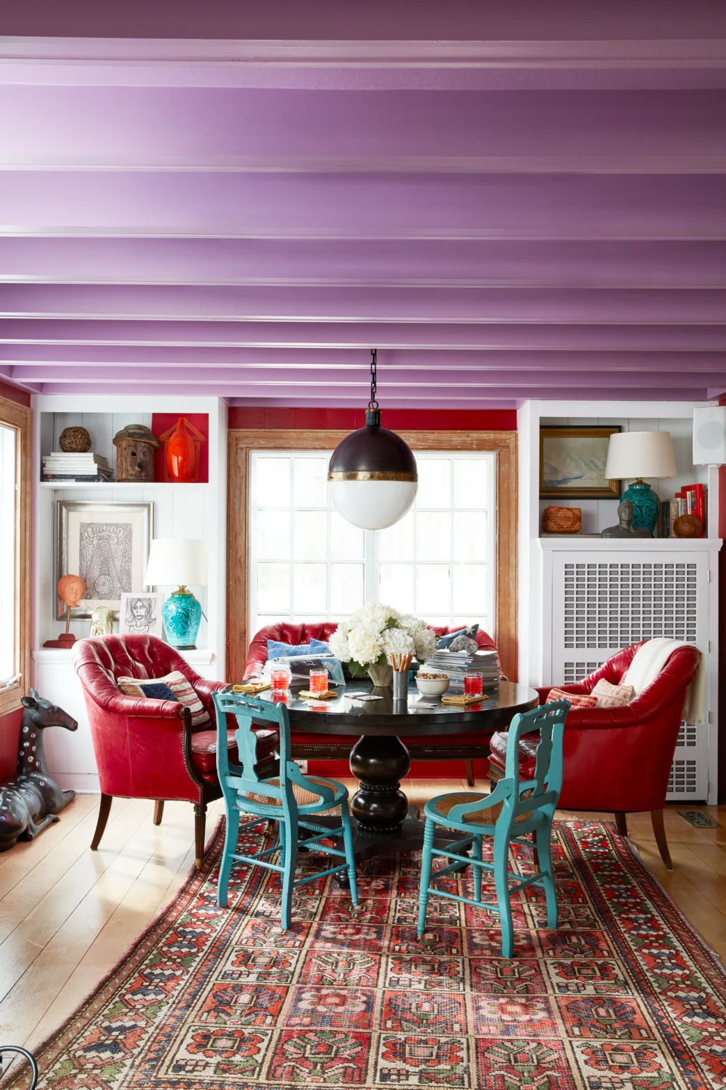 An eclectic dining room features a painted purple beamed ceiling, a round black table, red leather armchairs, and teal painted chairs.