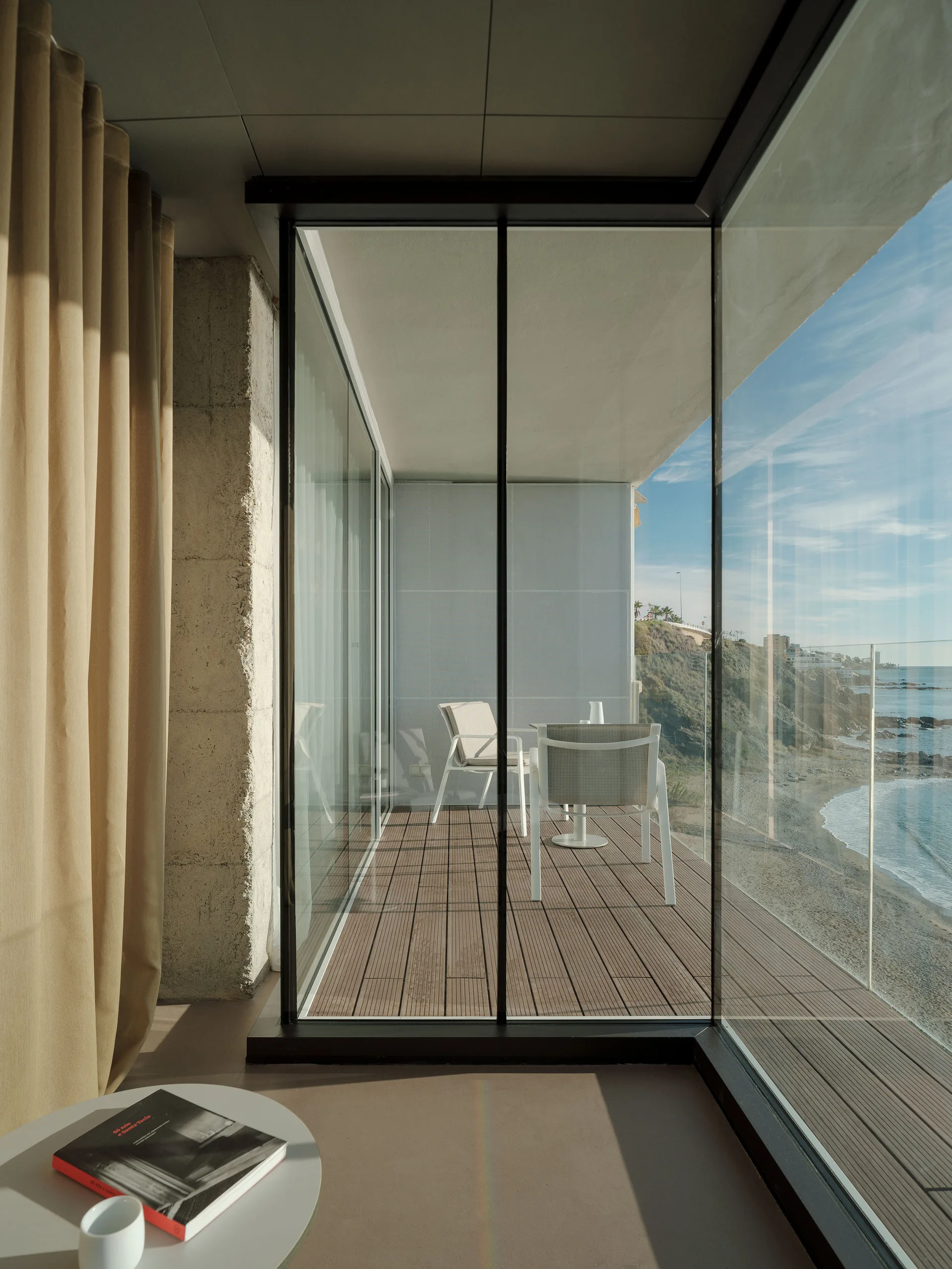 An indoor room with beige curtains looking through floor-to-ceiling glass windows onto a wood-planked terrace with outdoor seating overlooking the ocean.