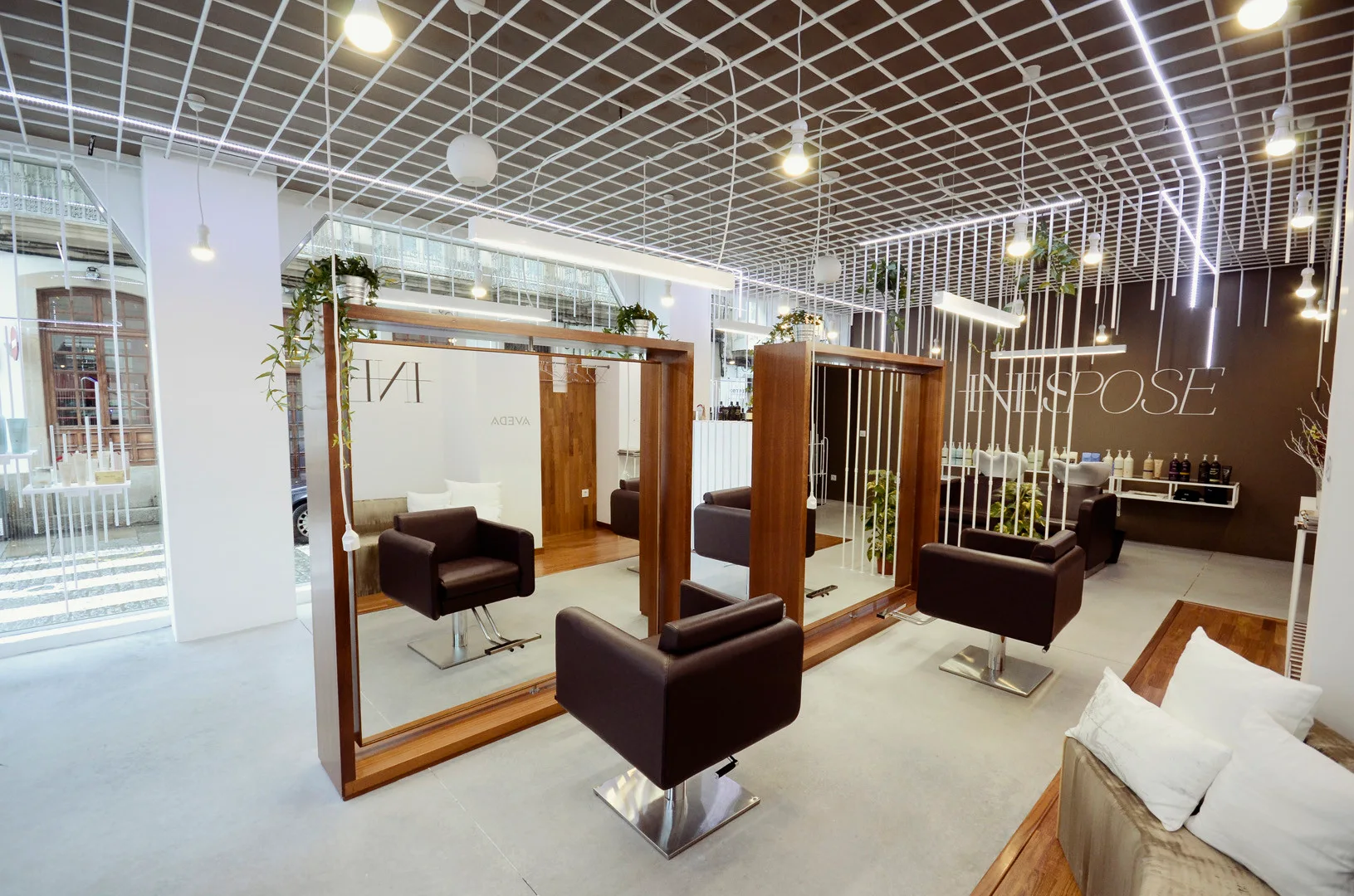 A modern salon interior features dark brown styling chairs facing large wood-framed mirrors beneath a white structural ceiling grid.