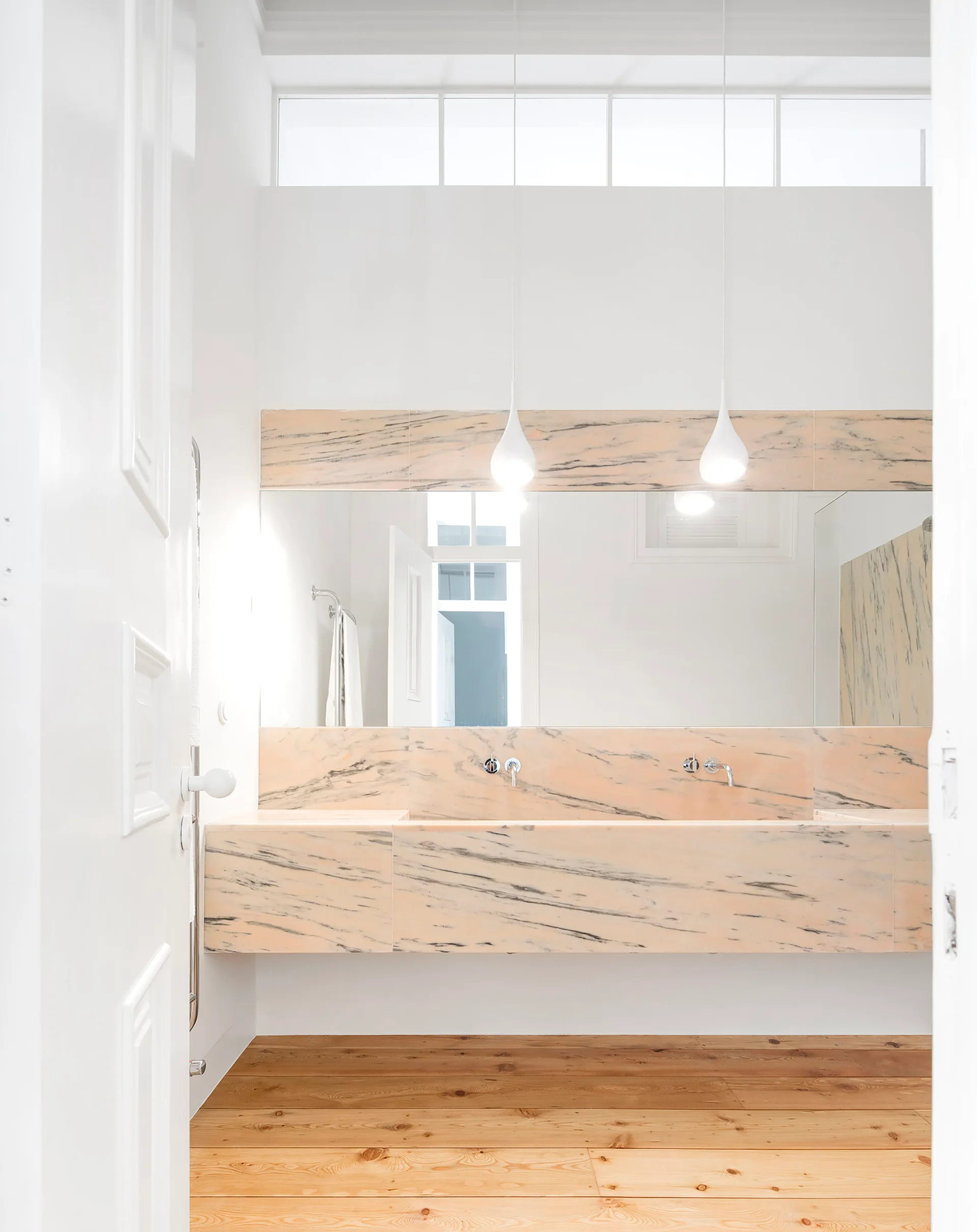 A minimalist bathroom features a floating pink-veined marble vanity beneath a large mirror, two white teardrop pendant lights, and light wood plank flooring.