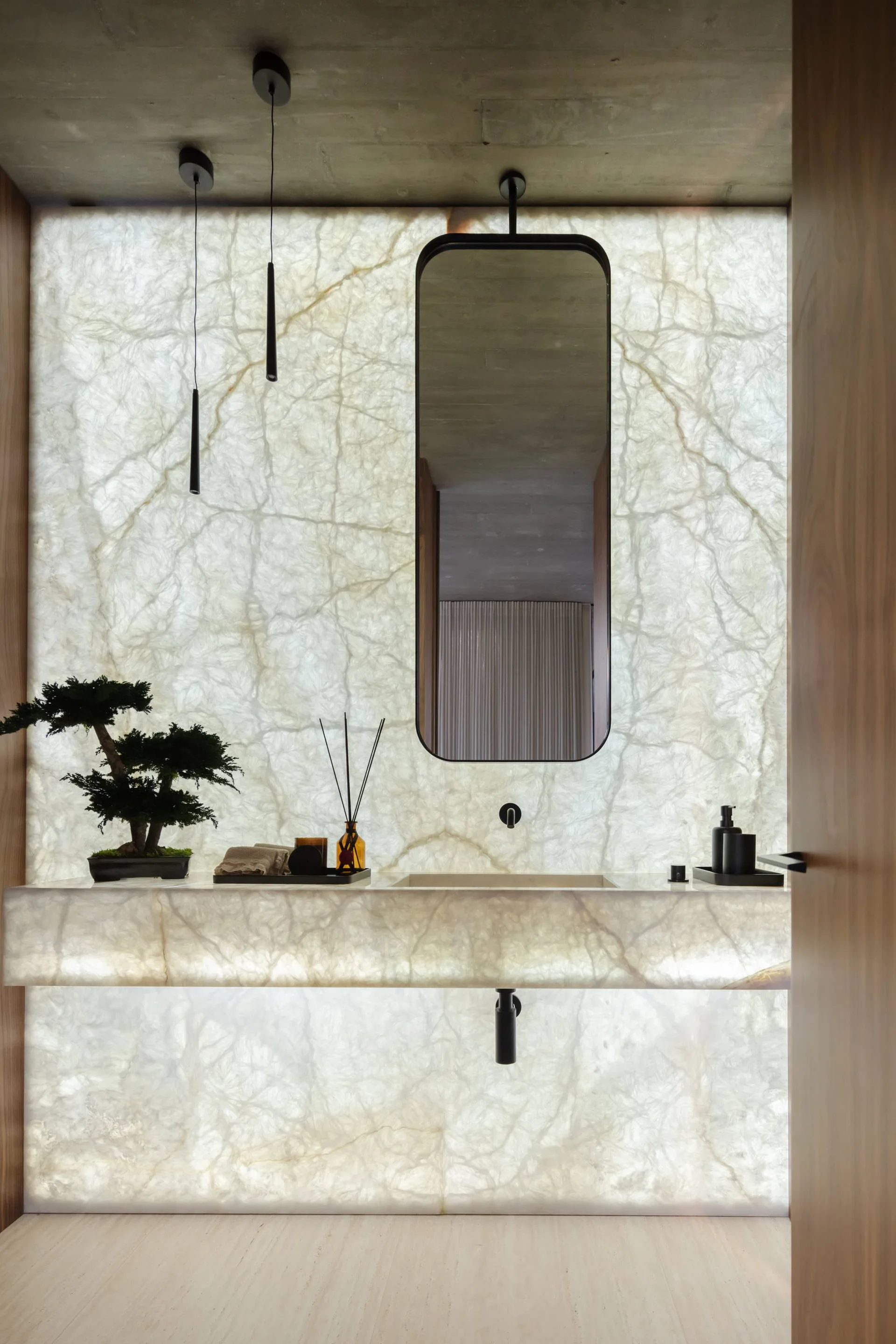 A modern bathroom vanity features a floating stone sink against an illuminated backlit translucent stone wall with a minimalist black framed mirror.