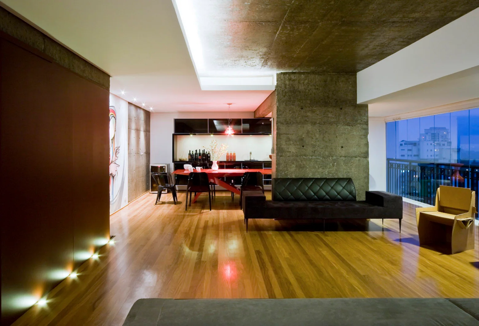 A modern open-plan loft features a black quilted sofa, an exposed concrete pillar, a red dining table, and polished wood flooring.