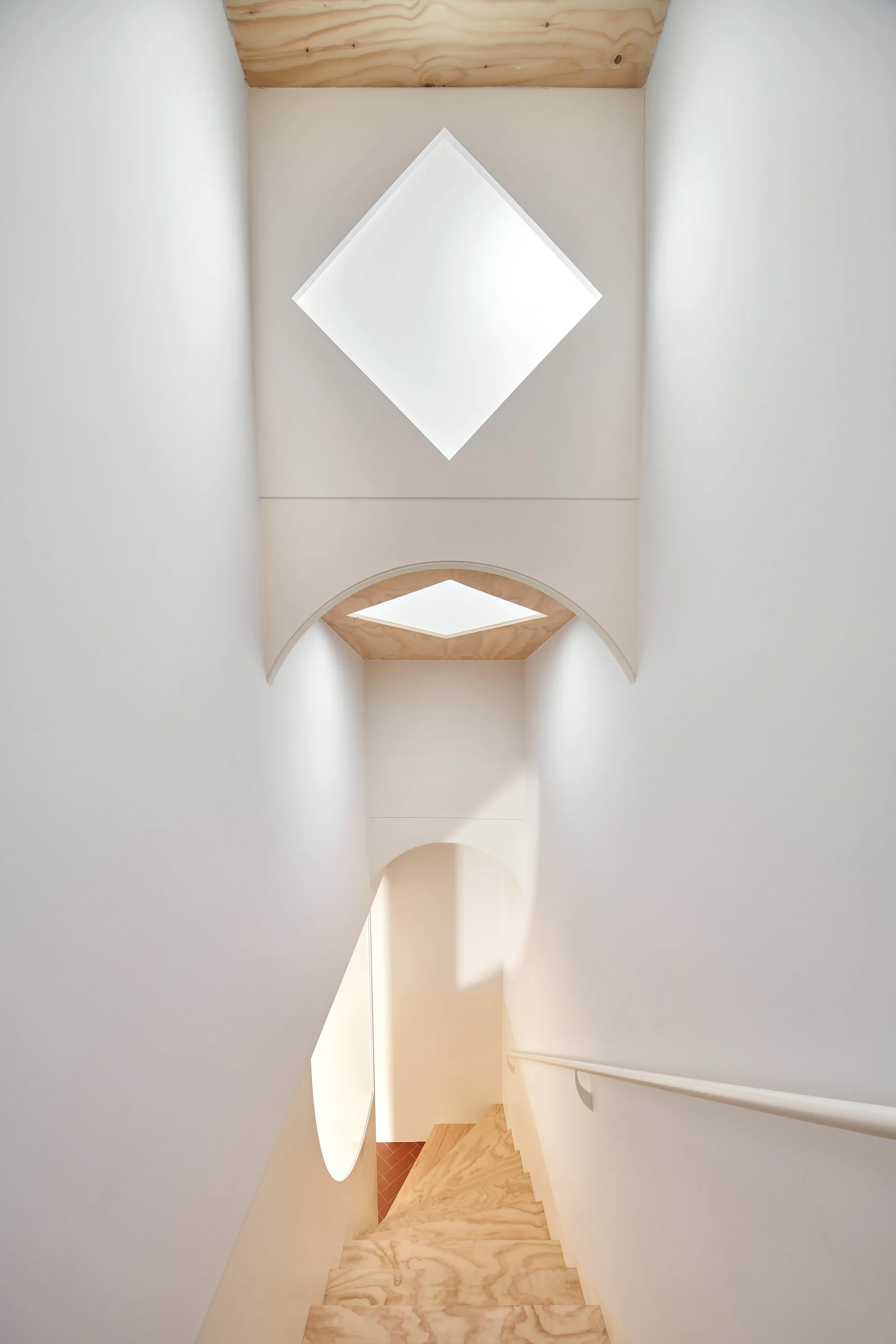 A downward view of a white minimalist staircase featuring light plywood steps, geometric diamond skylights, and clean arched overhead structures.