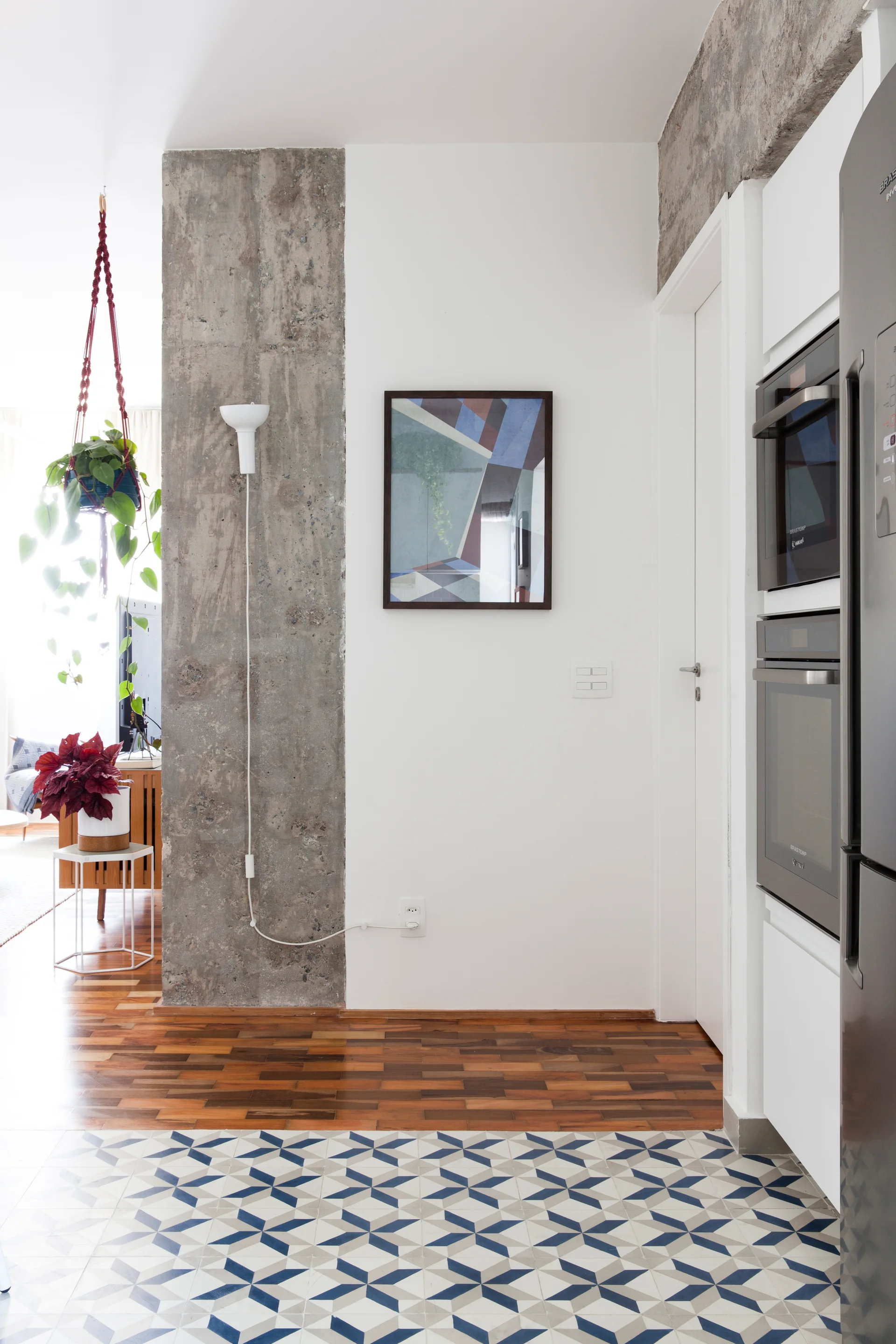 An interior view shows a transition from wood flooring to blue and white geometric tile, next to an exposed concrete column and white kitchen cabinetry.
