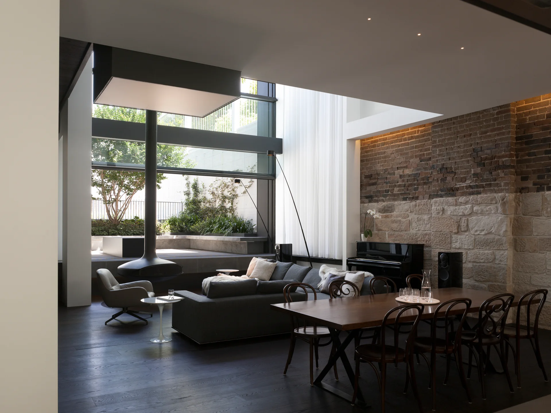 An open-plan living and dining space features dark wood floors, a grey sectional, a hanging black fireplace, and a textured brick and stone wall.