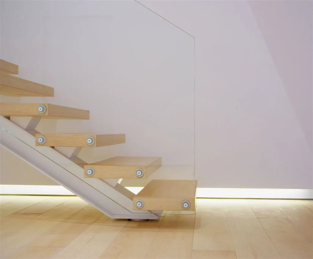 A modern staircase featuring pale wood floating treads with round metal mounts, a clear glass balustrade, and glowing baseboard strip lighting.
