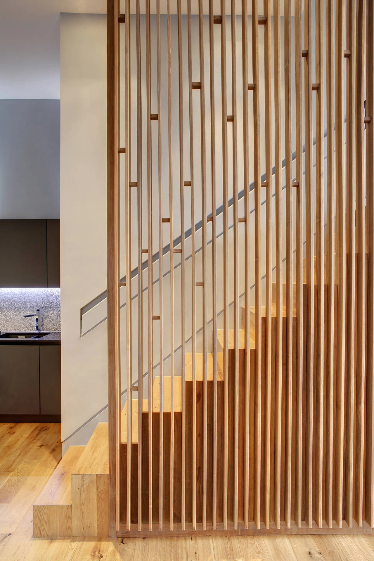 A modern wooden staircase featuring a tall partition made of vertical timber slats, with a kitchen visible in the background.