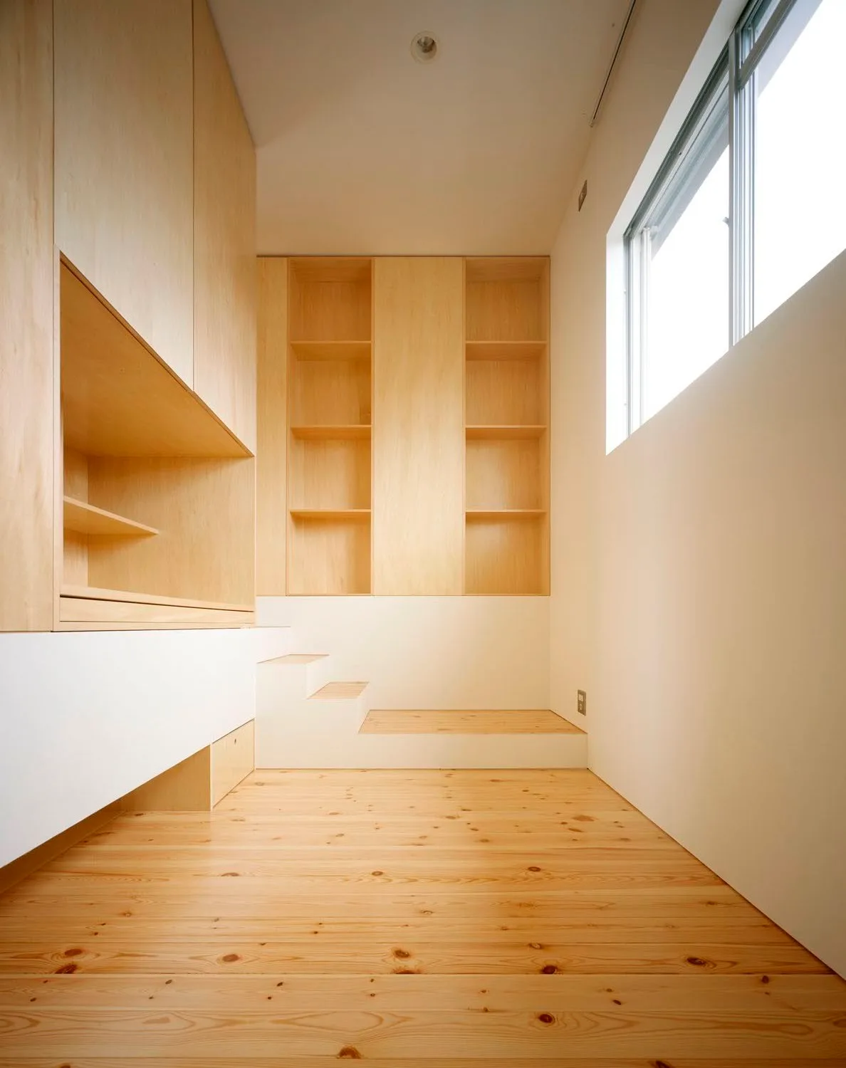 A narrow room features minimalist light wood built-in cabinets, recessed bookshelves, and small stepped platforms against white walls.