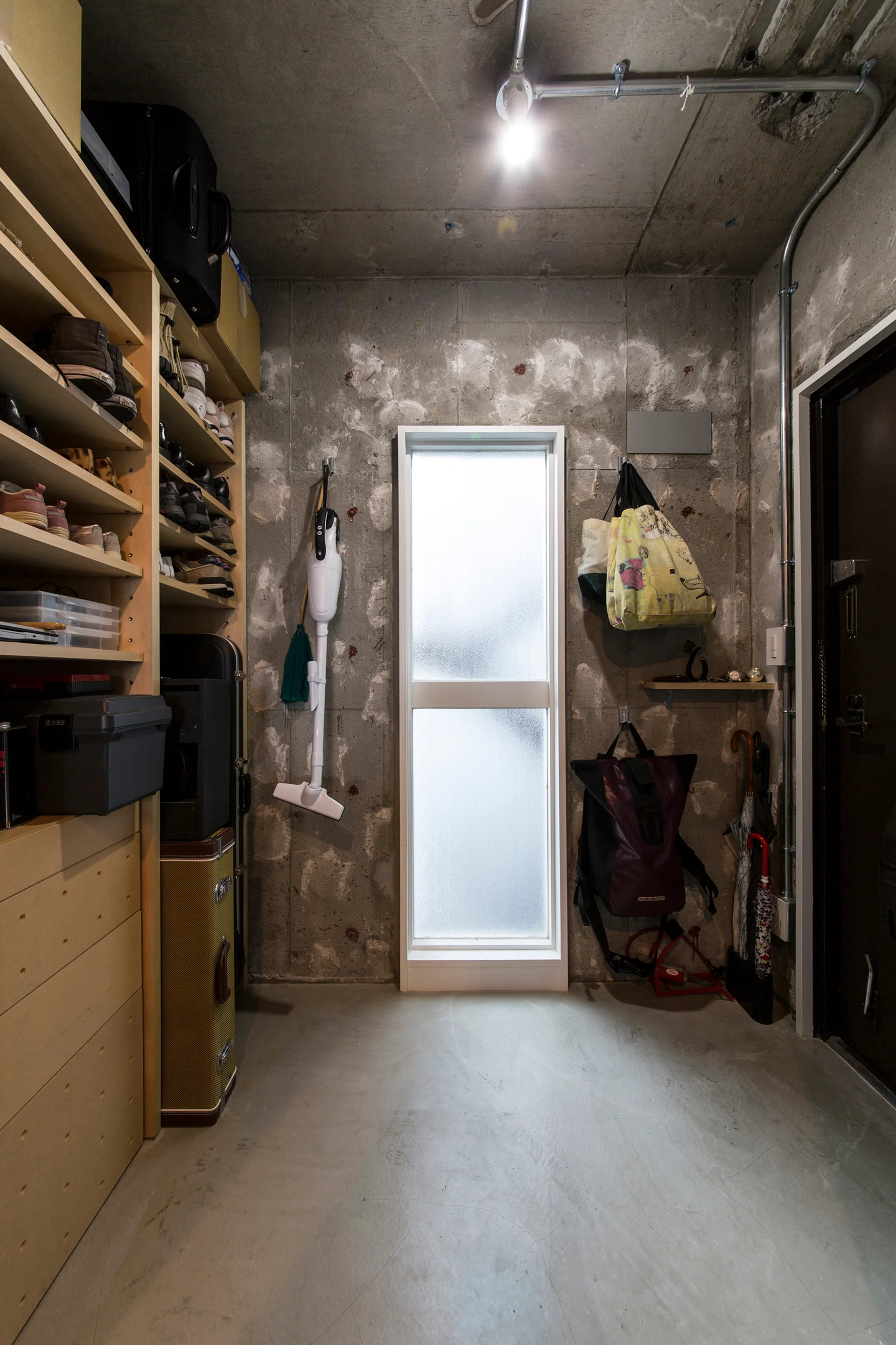 An industrial mudroom features raw concrete walls, a tall wooden storage shelving unit, and a slim frosted glass window in the center.