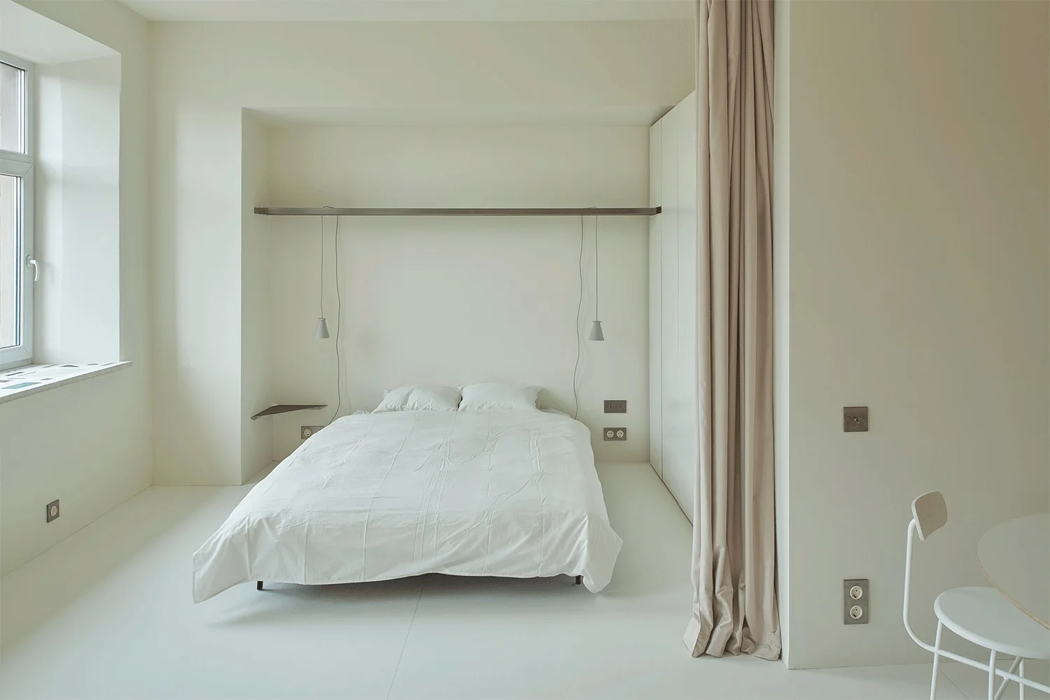 A minimalist monochromatic white bedroom featuring a bed in a recess beneath a thin metal shelf and a soft beige curtain partition.