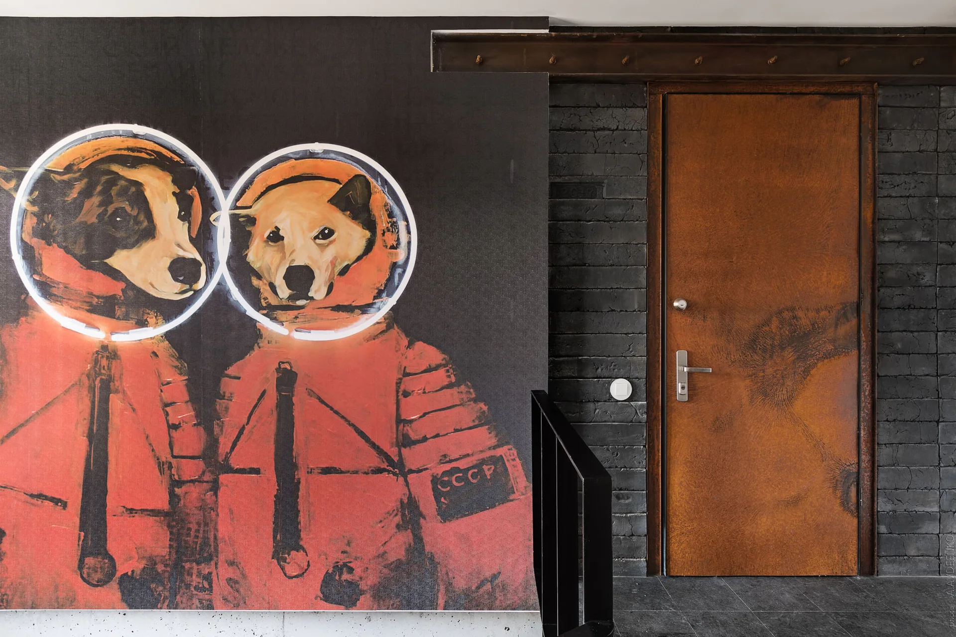A rust-colored metal door stands next to a dark painted brick wall featuring a large mural of two dogs in orange space suits.
